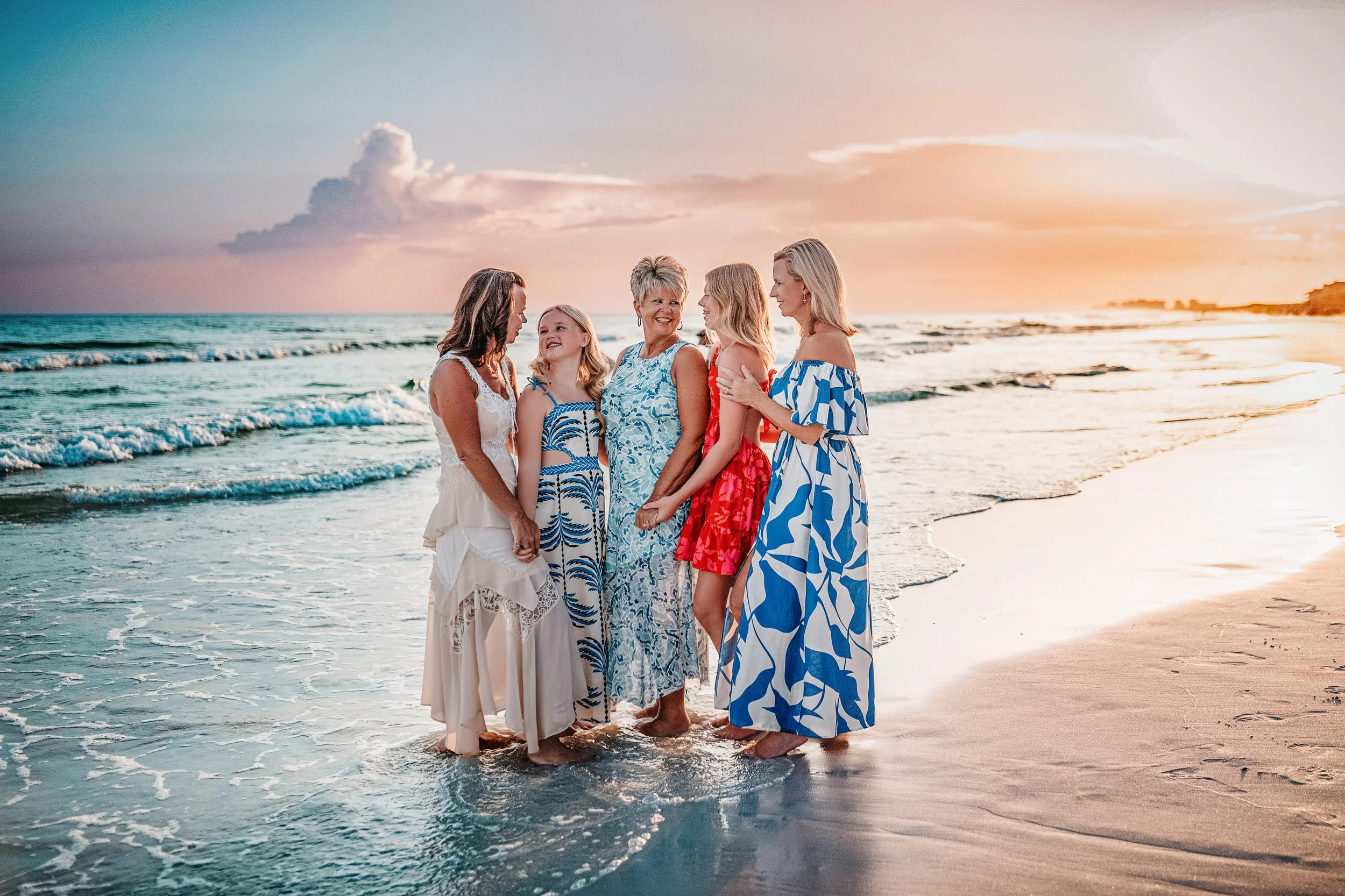 Five women and girls sharing a candid moment together in the shallow Gulf water during a colorful sunset family beach session along Florida’s Emerald Coast.