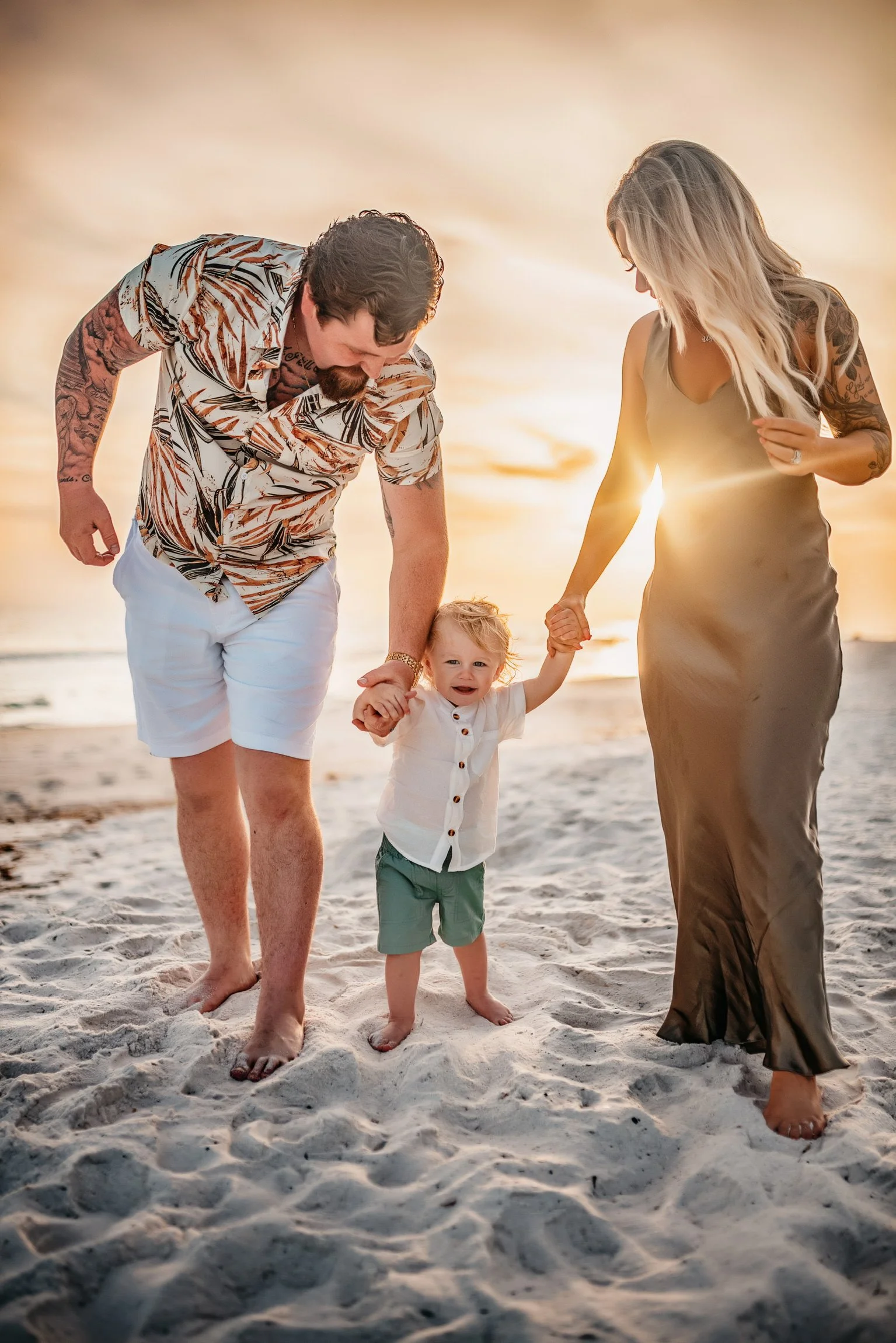 A family of three holding hands and walking on the beach at sunset, with the child in the middle. The father is wearing a tropical shirt and white shorts, the mother is in a gold satin dress, and the child has a white shirt and green shorts.