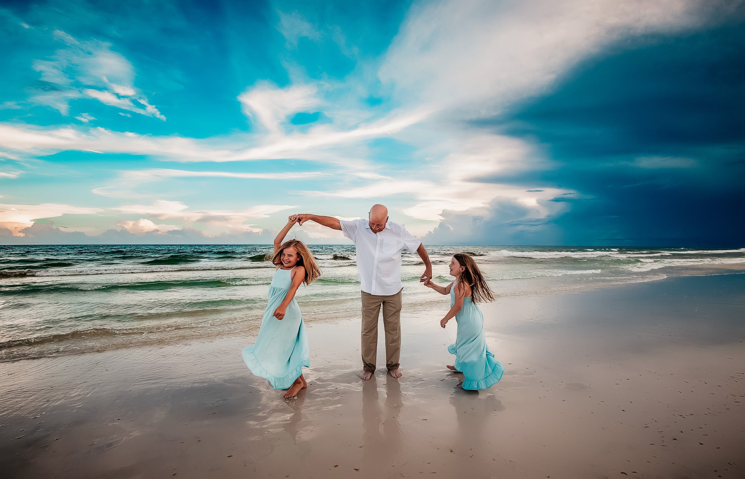 A man and two young girls dancing on a beach during a storm, with dramatic clouds and waves in the background, all wearing light blue dresses.