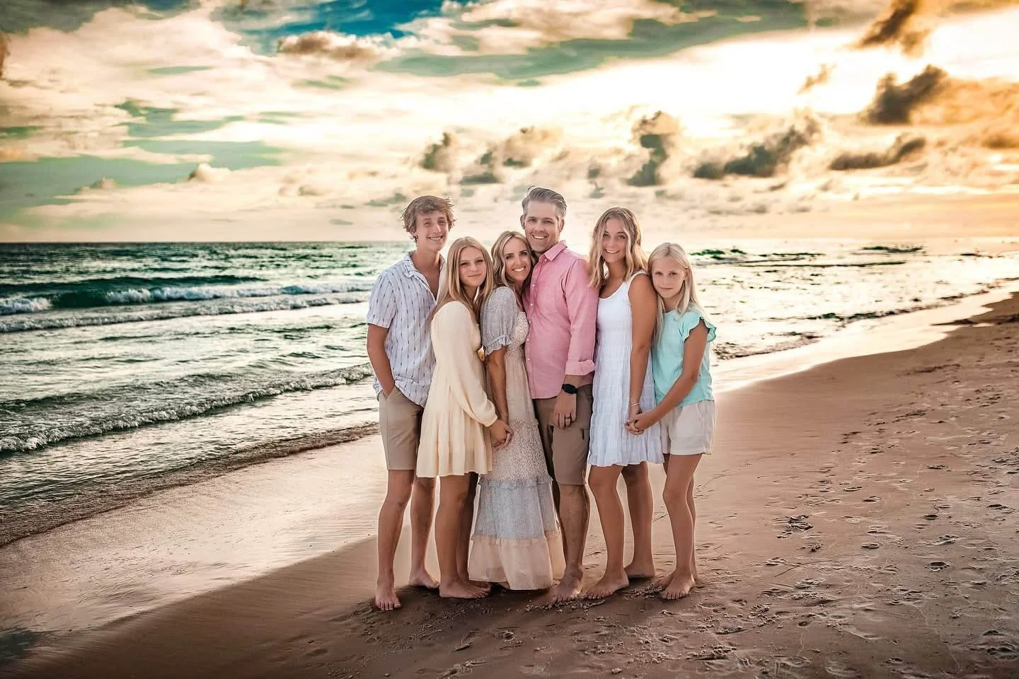 A family of seven, including adults and children, stands together on a beach at sunset, smiling and facing the camera.