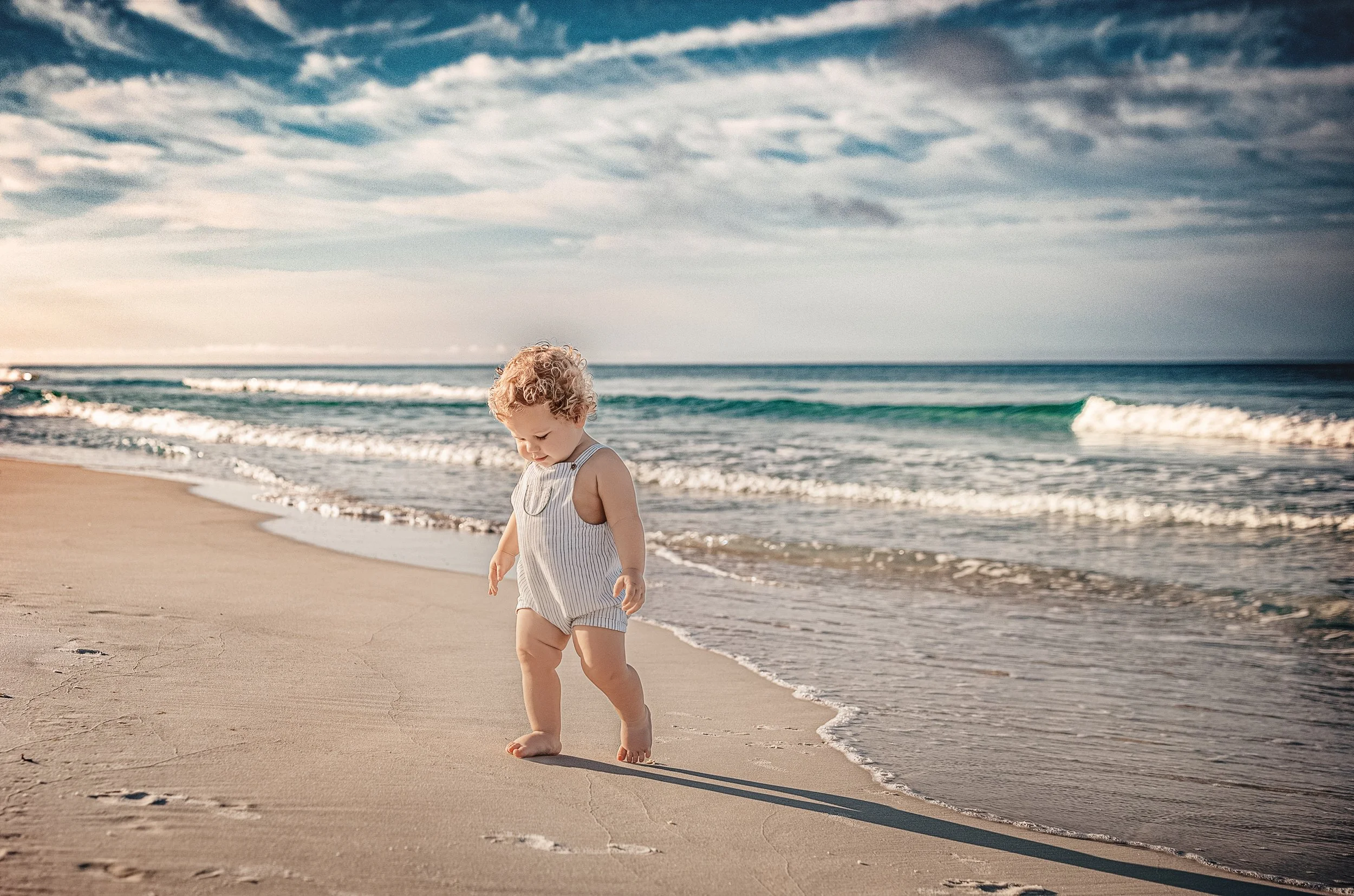 Curly-haired toddler in a striped romper walking barefoot along the shoreline at 30A Florida, emerald green Gulf waves rolling in behind him during a morning family beach session