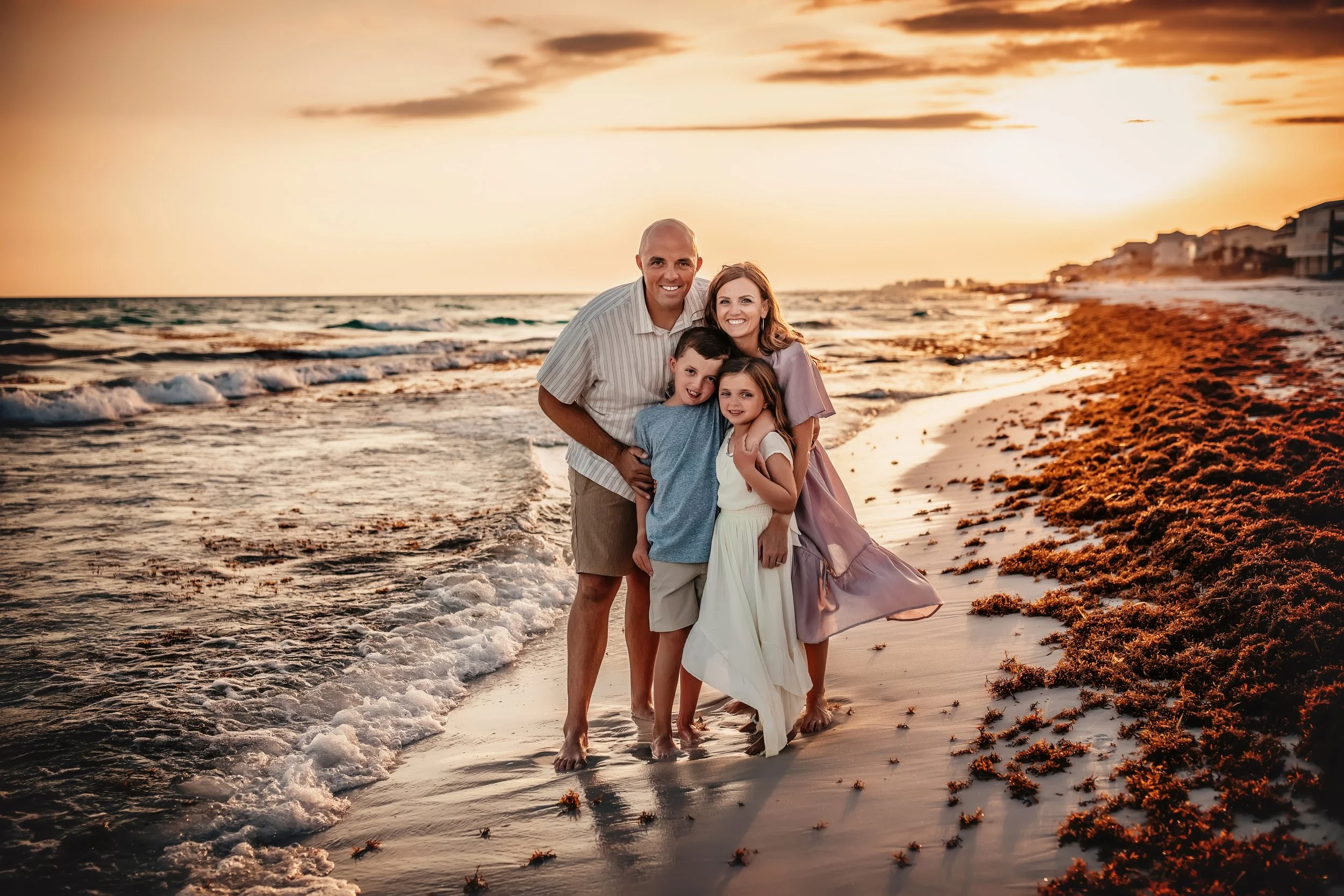 A mother and father embracing their little boy and little girl on the shore looking at the camera smiling during their sunset beach session