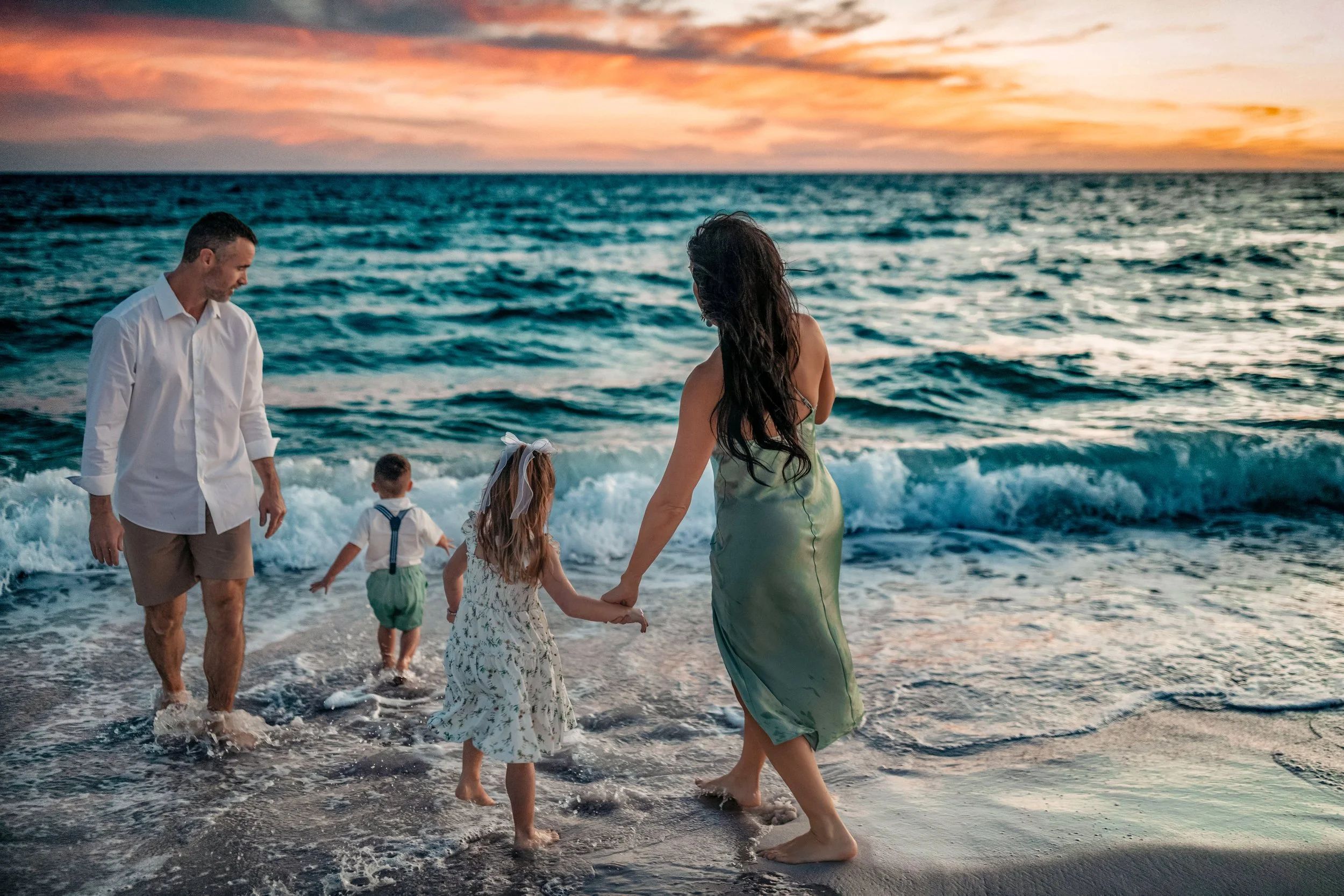 Full body shot of a family of three walking hand-in-hand through the shallow surf at Santa Rosa Beach, FL, featuring a boho-style green dress.