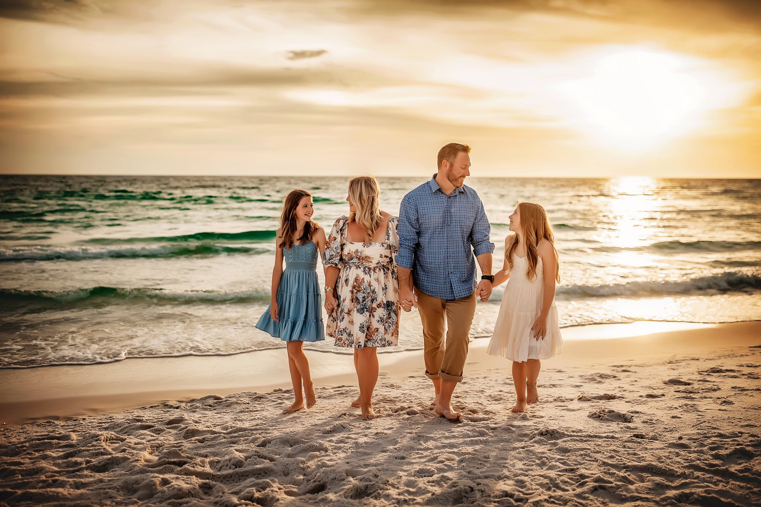 A family of four walking on the beach at sunset, holding hands and smiling at each other.