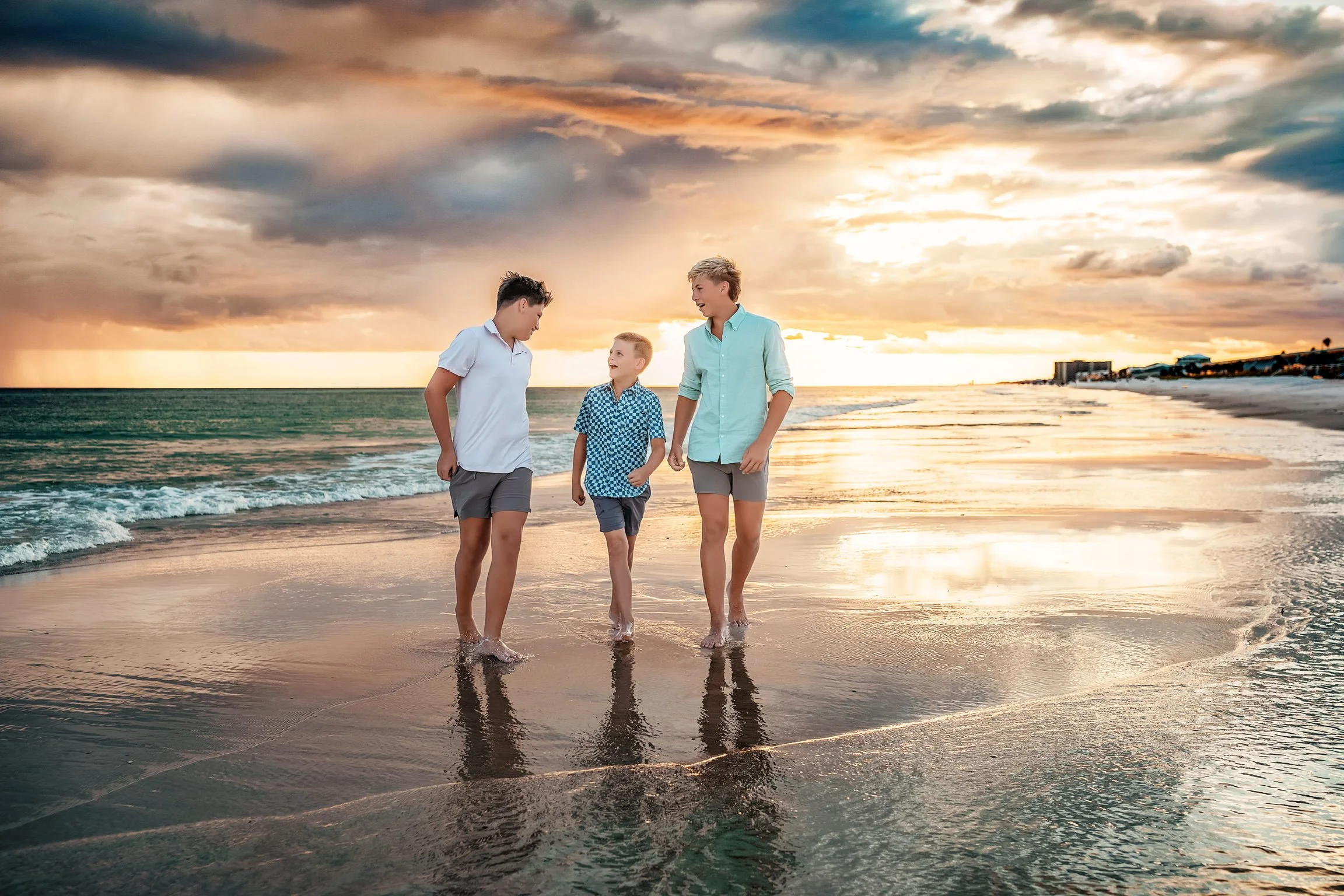 Three boys walking on the beach at sunset, holding hands and talking, with ocean waves and cloudy sky in the background.