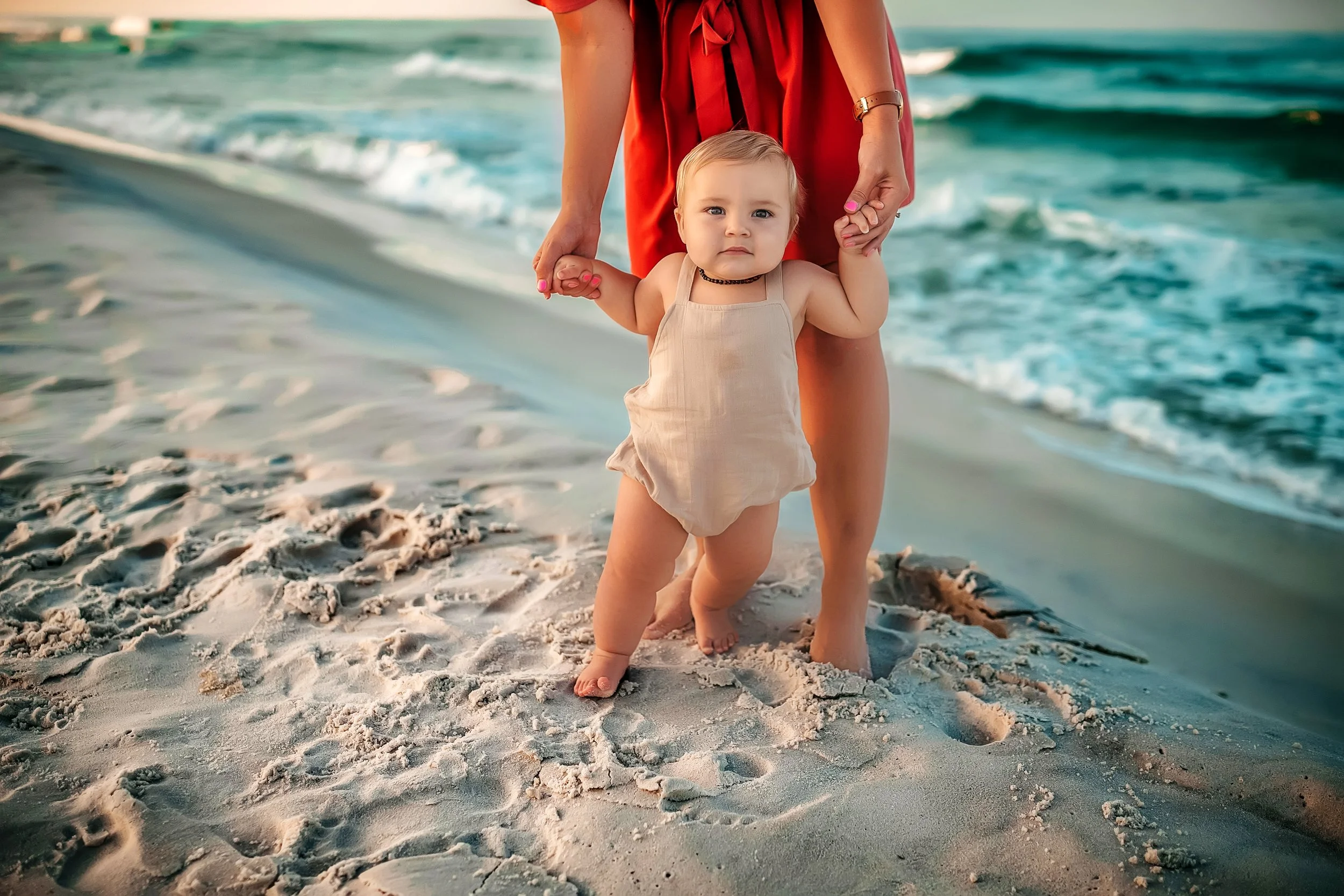 Baby holding hands with parent while learning to walk on the shoreline during a natural beach photography session on 30A.