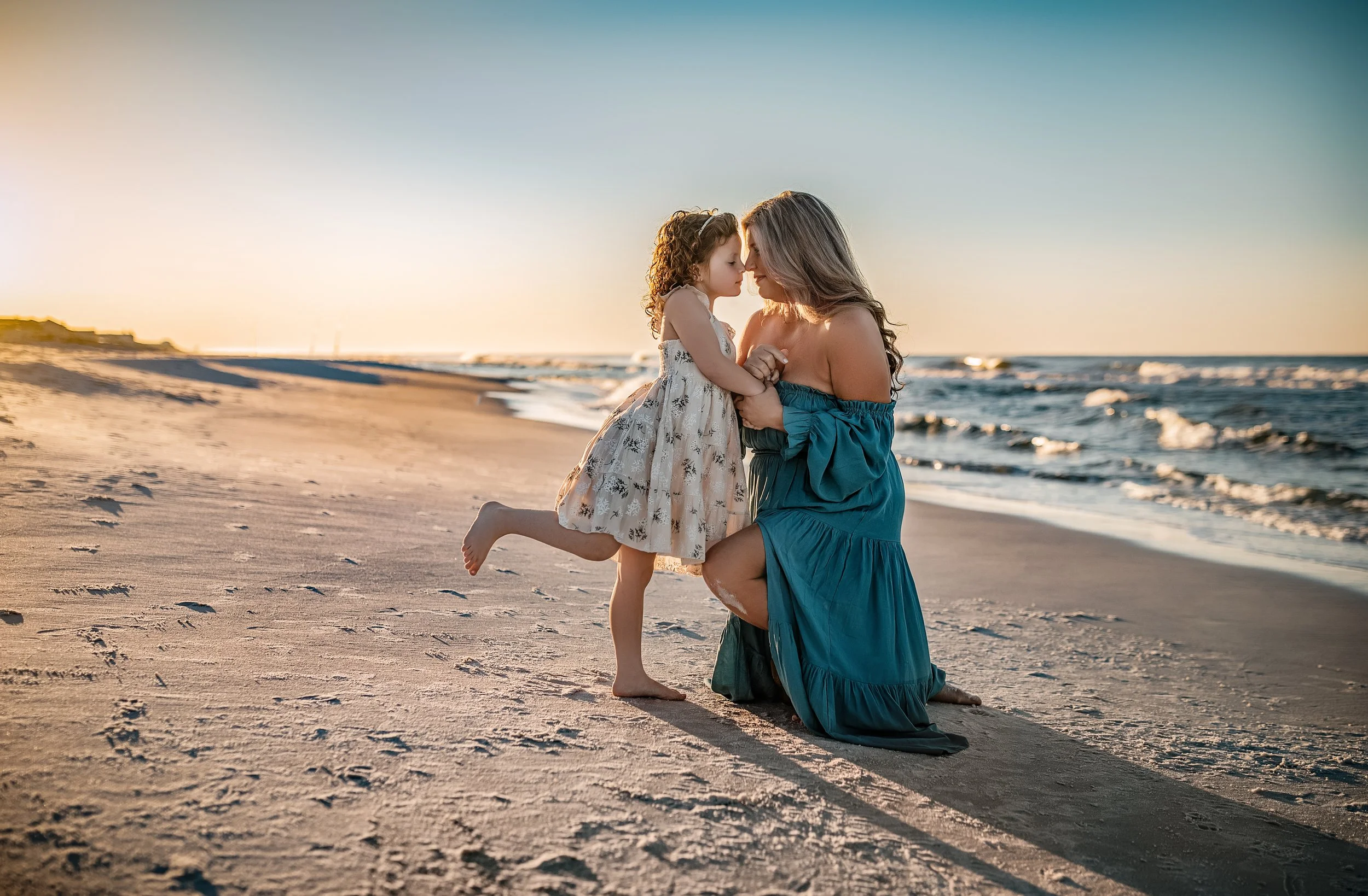 A mother in a teal off-the-shoulder dress sharing a tender moment with her young daughter on the sand at Santa Rosa Beach, Florida during golden hour.