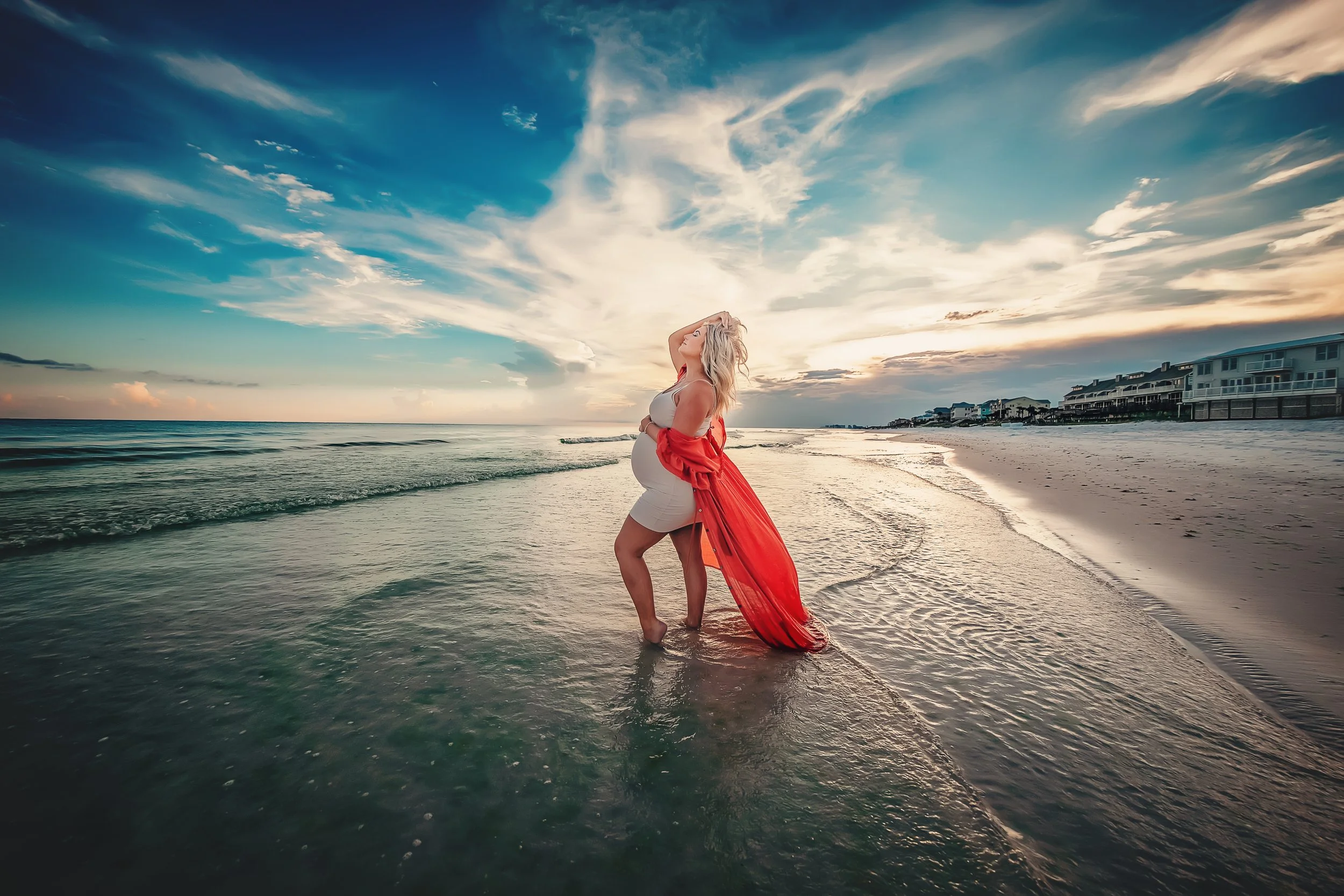 Editorial maternity portrait of a woman in a white dress and flowing red silk wrap standing in the Gulf of Mexico waters at Santa Rosa Beach under a dramatic blue sky.