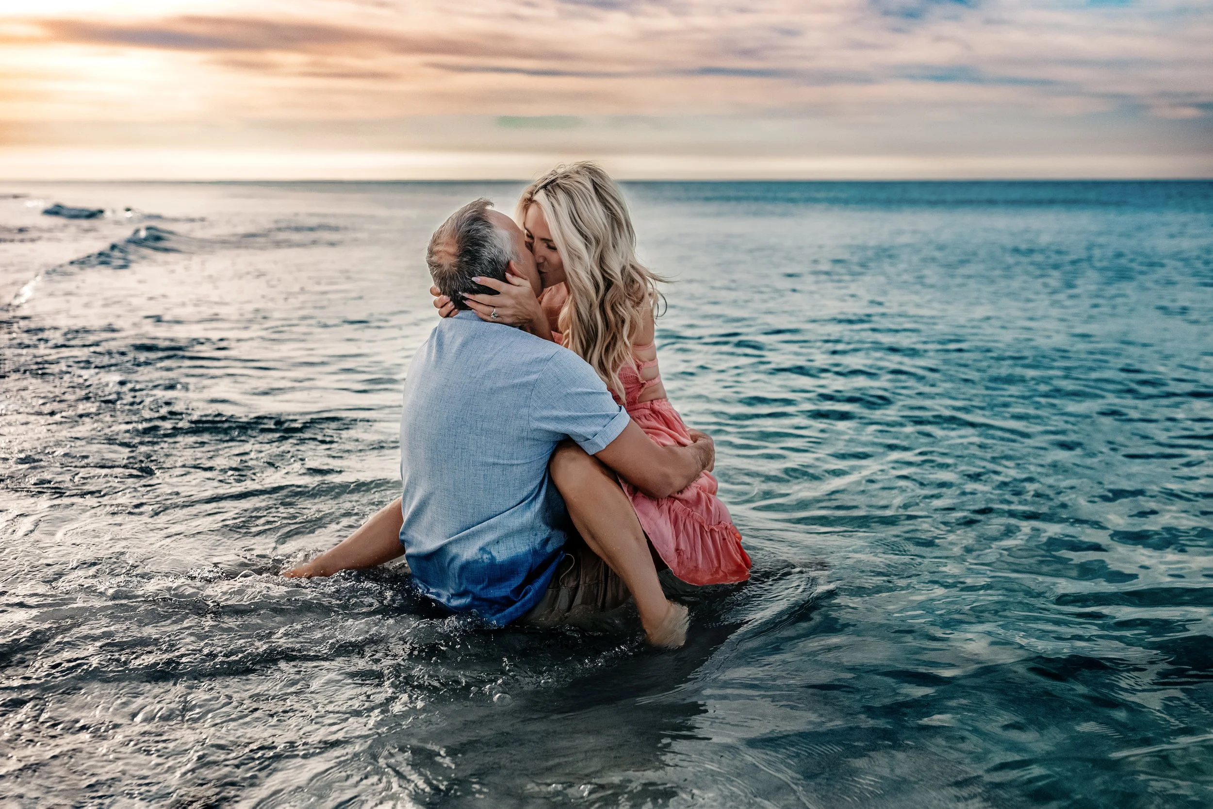 Couple sitting in ocean water at sunset during 30A beach photo session — woman in coral pink dress, man in blue chambray shirt — Gulf Coast Florida photographer