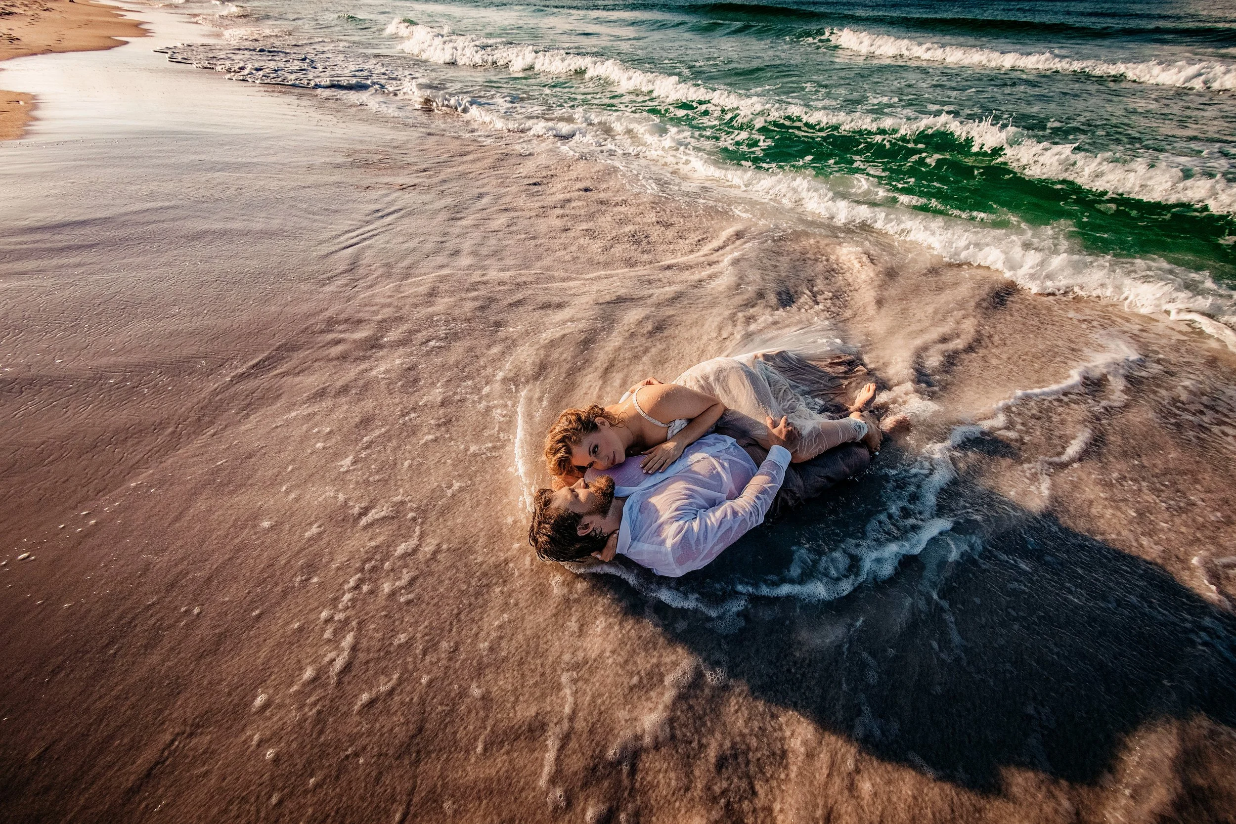 Bride and groom lying together in shoreline foam during adventurous 30A  beach wedding