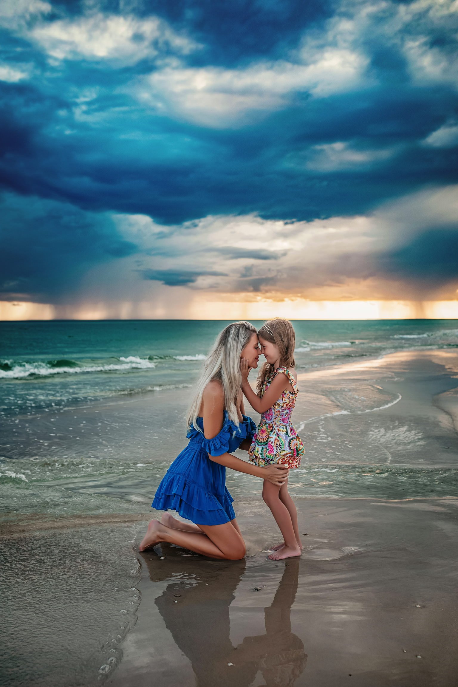 A woman kneeling in the sand, touching foreheads with a young girl on a beach during a stormy sunset.