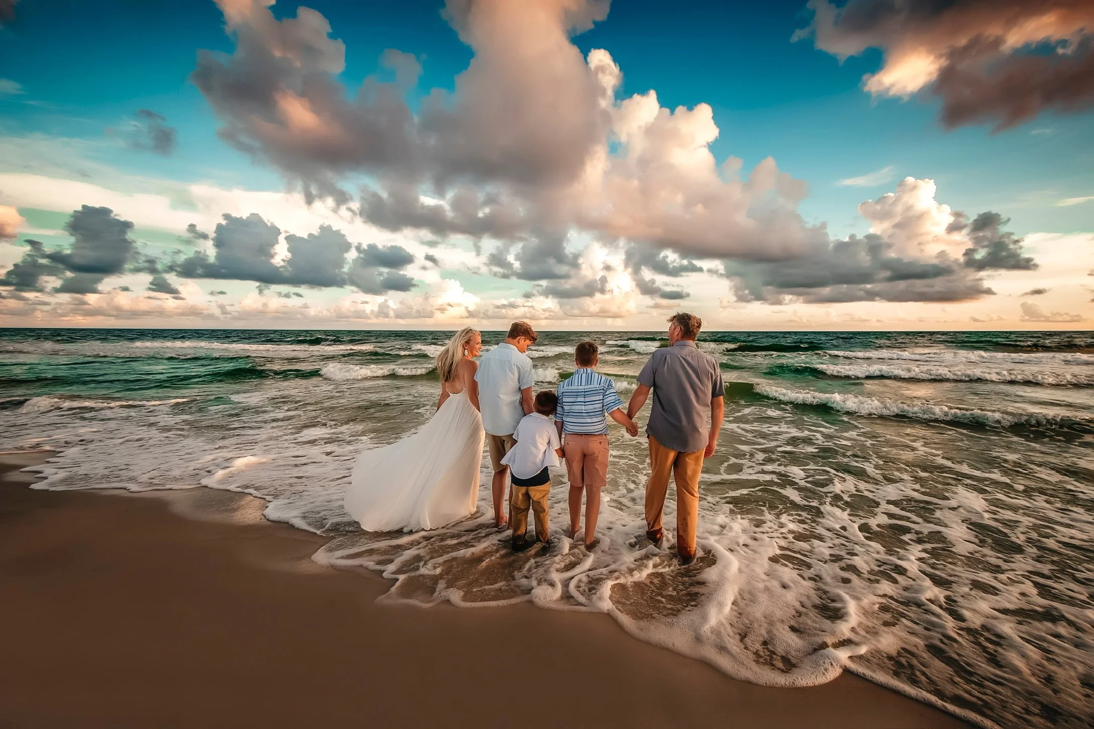 A group of six people, including a bride in a white wedding dress, standing on a sandy beach at sunset, holding hands and looking out at the ocean with clouds in the sky.
