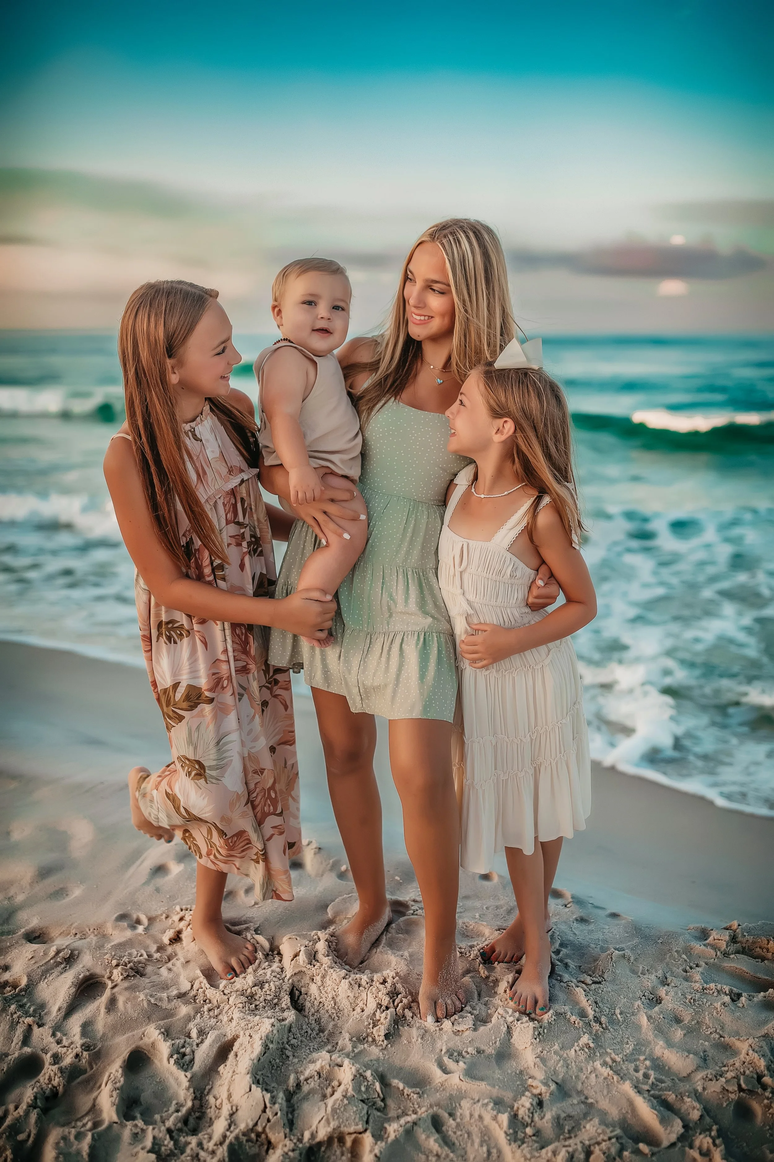 Sisters smiling and holding a baby during a golden hour beach photo session on 30A with soft neutral outfits.