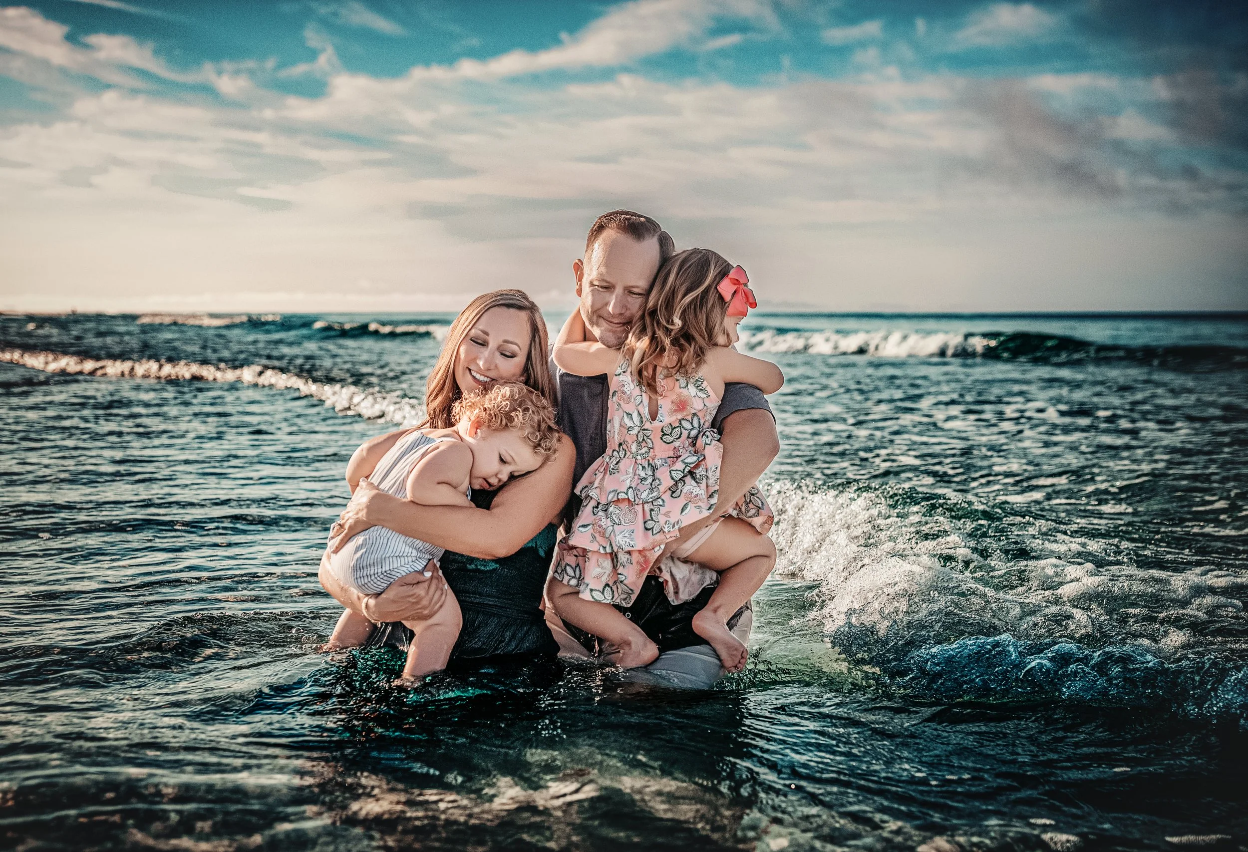 Family of four sitting together in the Gulf of Mexico waves on 30A, mom and dad each holding a young child, laughing and splashing during a beach photography session on the Emerald Coast of Florida
