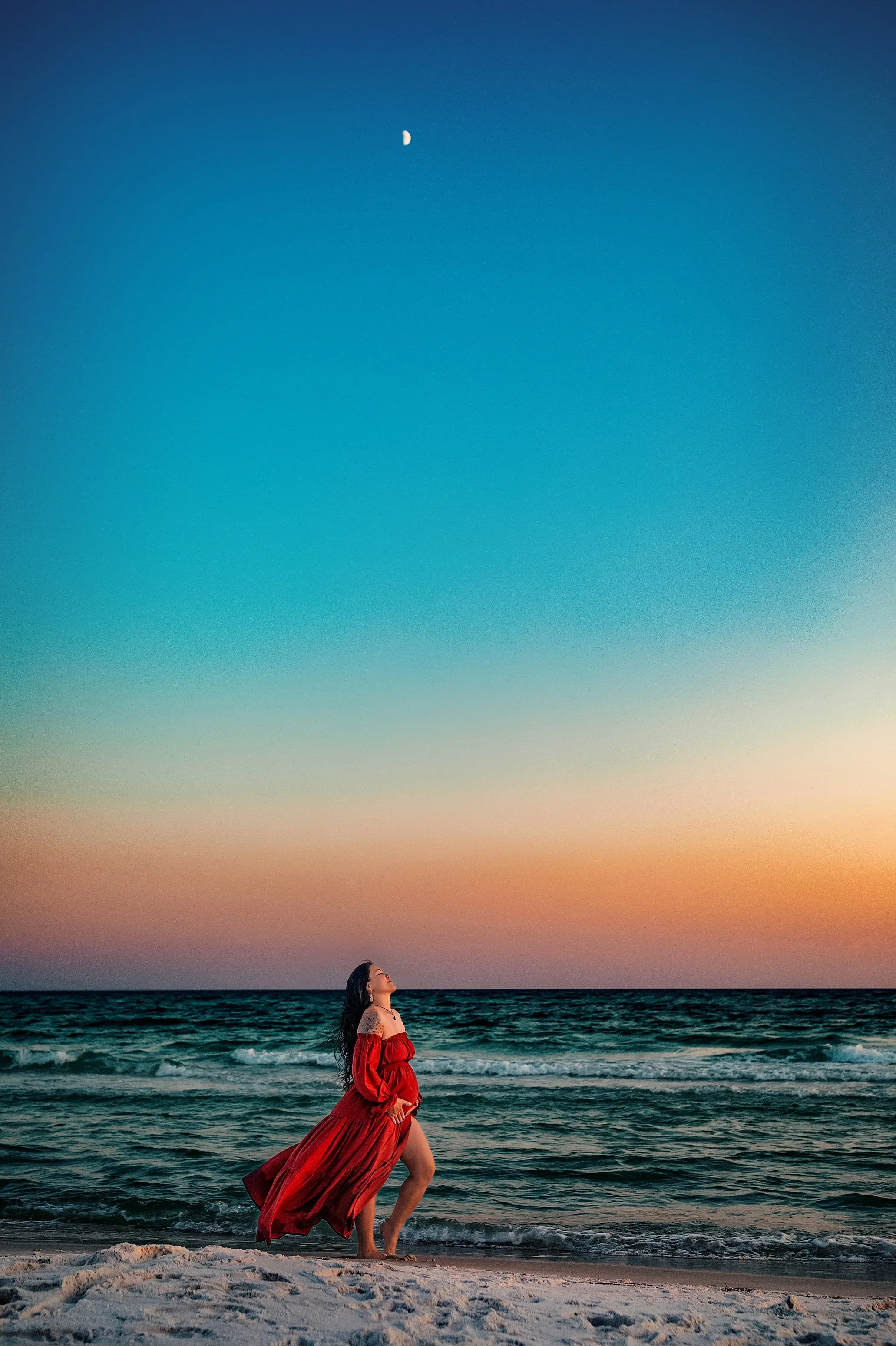 Expectant mother in a flowing red gown posing against a vibrant sunset sky on a 30A beach