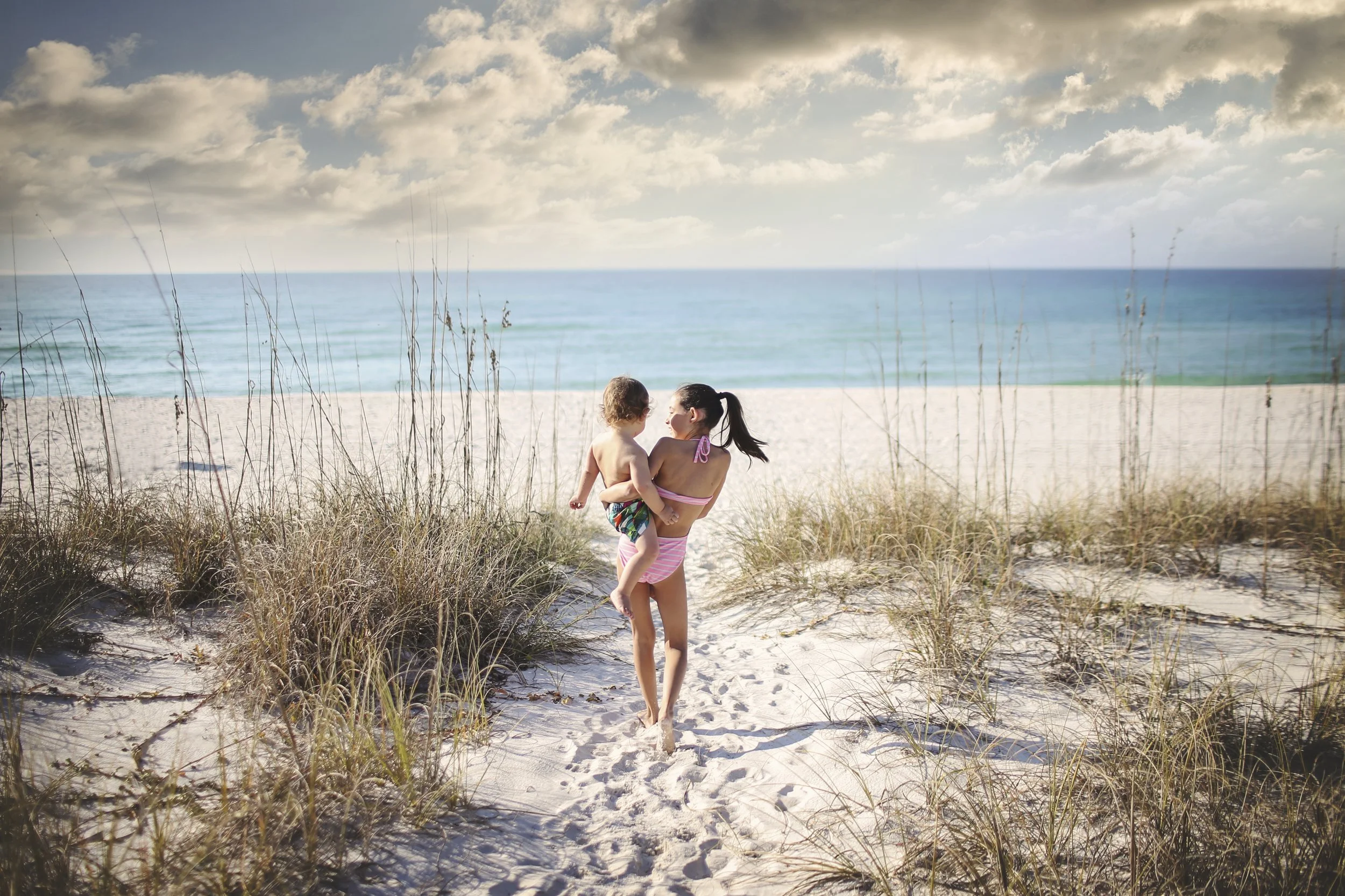 A woman holding a child walking towards the beach through the sand dunes with grassy plants, ocean and cloudy sky in the background.
