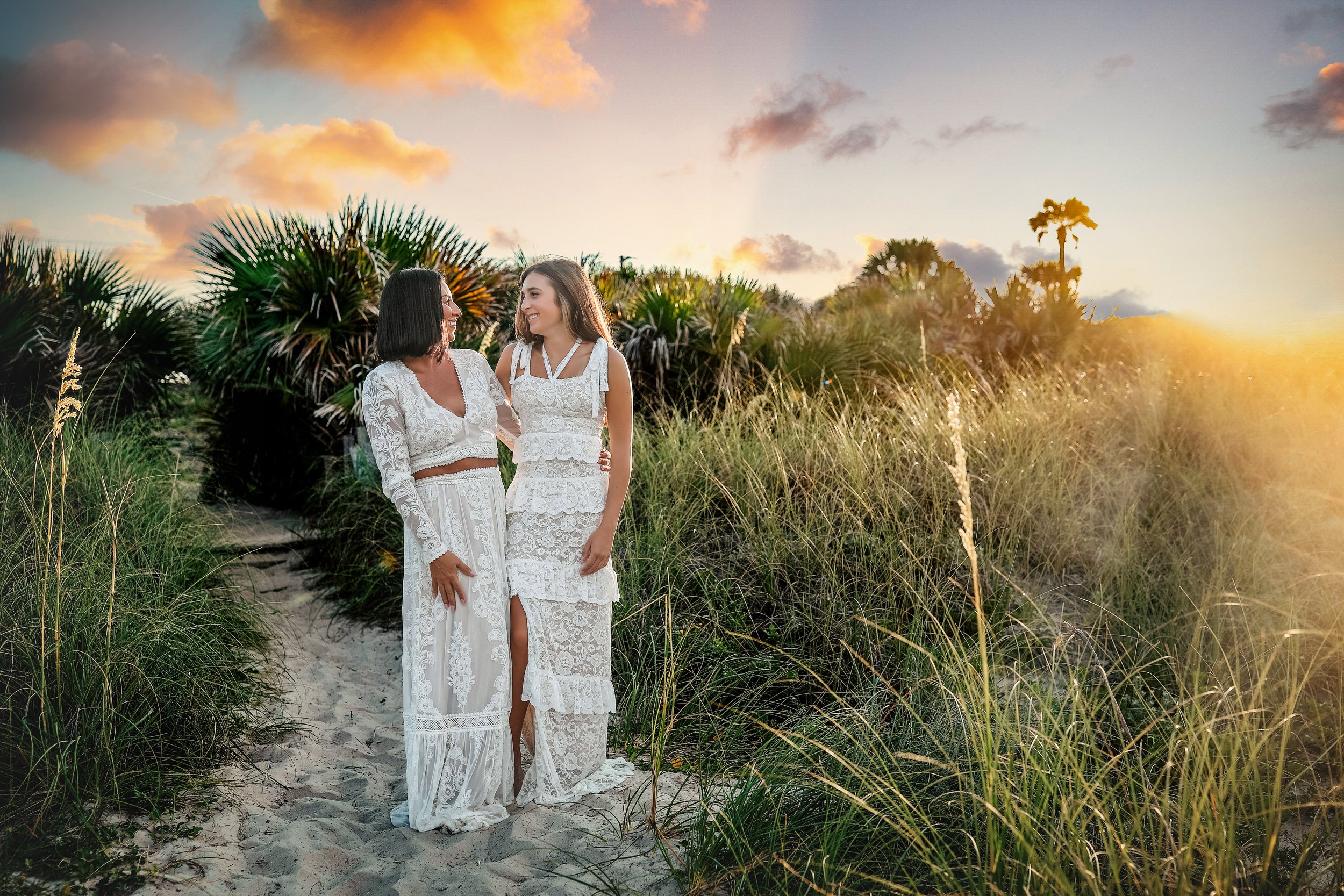 Two women in white lace dresses standing on a sandy path amidst tall grass and tropical plants, smiling at each other during sunset.