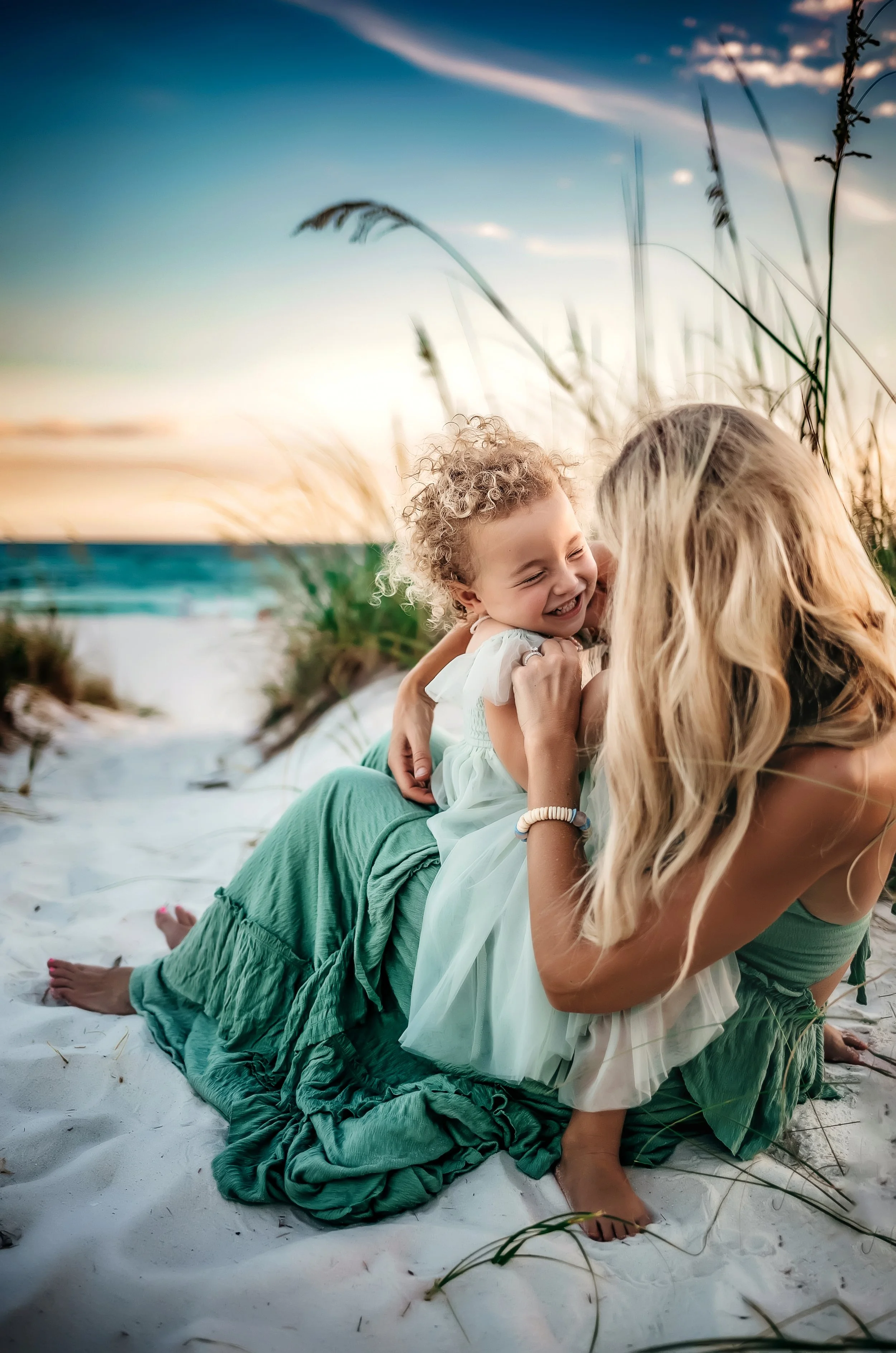 30A beach photographer capturing mother and daughter moment at sunset — woman lying in sand with girl sitting on her back laughing, beach grass and colorful Gulf Coast sky