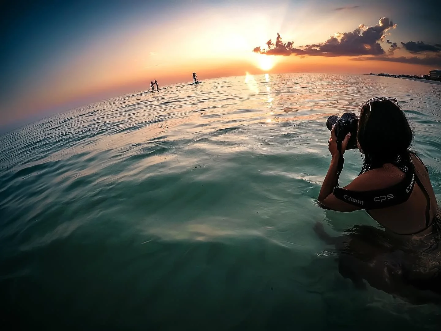 A woman taking a photograph with a camera while waist-deep in ocean water during sunset, with two paddleboarders and a city skyline in the distance.