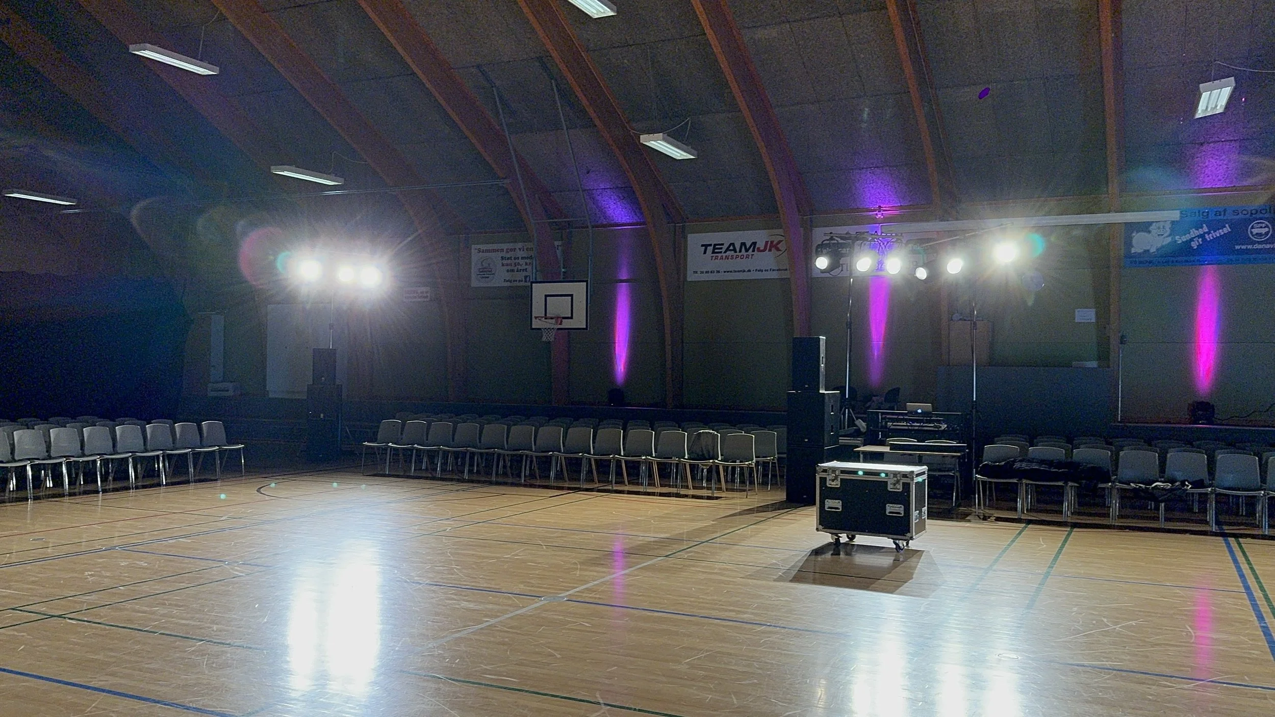 Indendørs basketballarena med tomt gulv, omringet af rækker af stole og lyd- og lysudstyr til en begivenhed, med belysning på væggene.