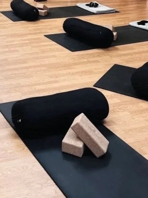 Yoga studio with black mats, bolsters, and bricks arranged on a wooden floor.