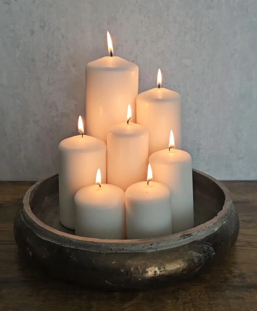 Seven lit white pillar candles arranged in a metal bowl on a wooden surface, against a plain wall.