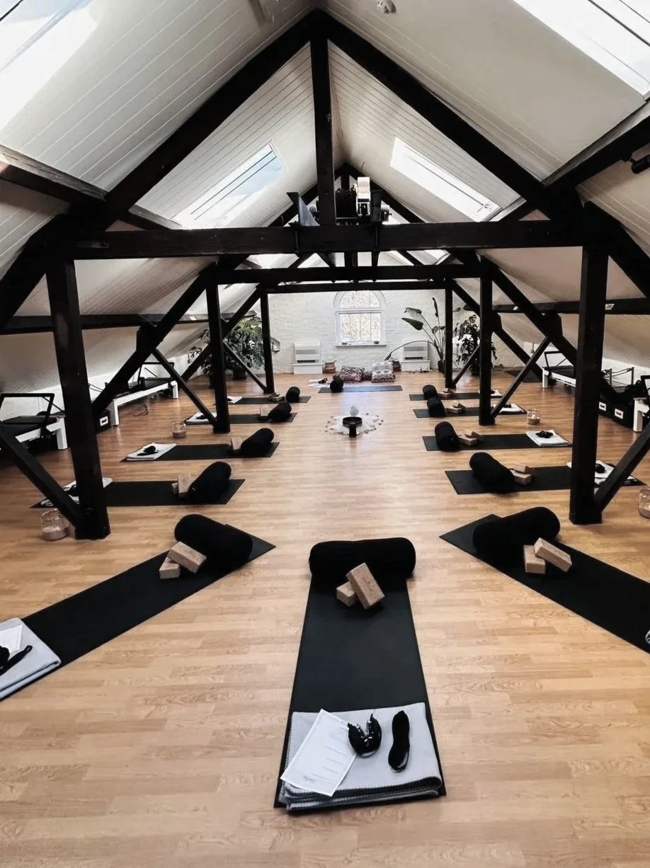 A spacious yoga studio with black mats, yoga blocks, and rolled towels arranged neatly on each mat. The room has a wooden floor, exposed dark beams overhead, and two large skylights providing natural light. Some plants and a window are visible at the back of the room.