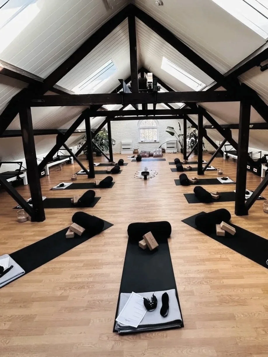 Yoga studio with black mats, yoga blocks, and bolsters arranged on a wooden floor in an attic space with skylight windows, plants, and a white brick wall.