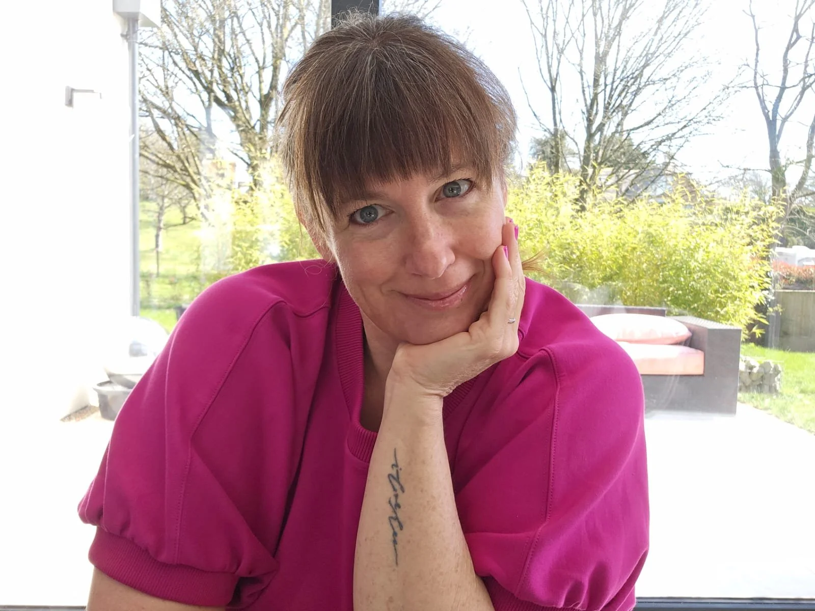 Melanie Cohen wearing a pink shirt, is resting her face on her hand and smiling slightly, sitting in front of a window with a garden view of trees and shrubs.