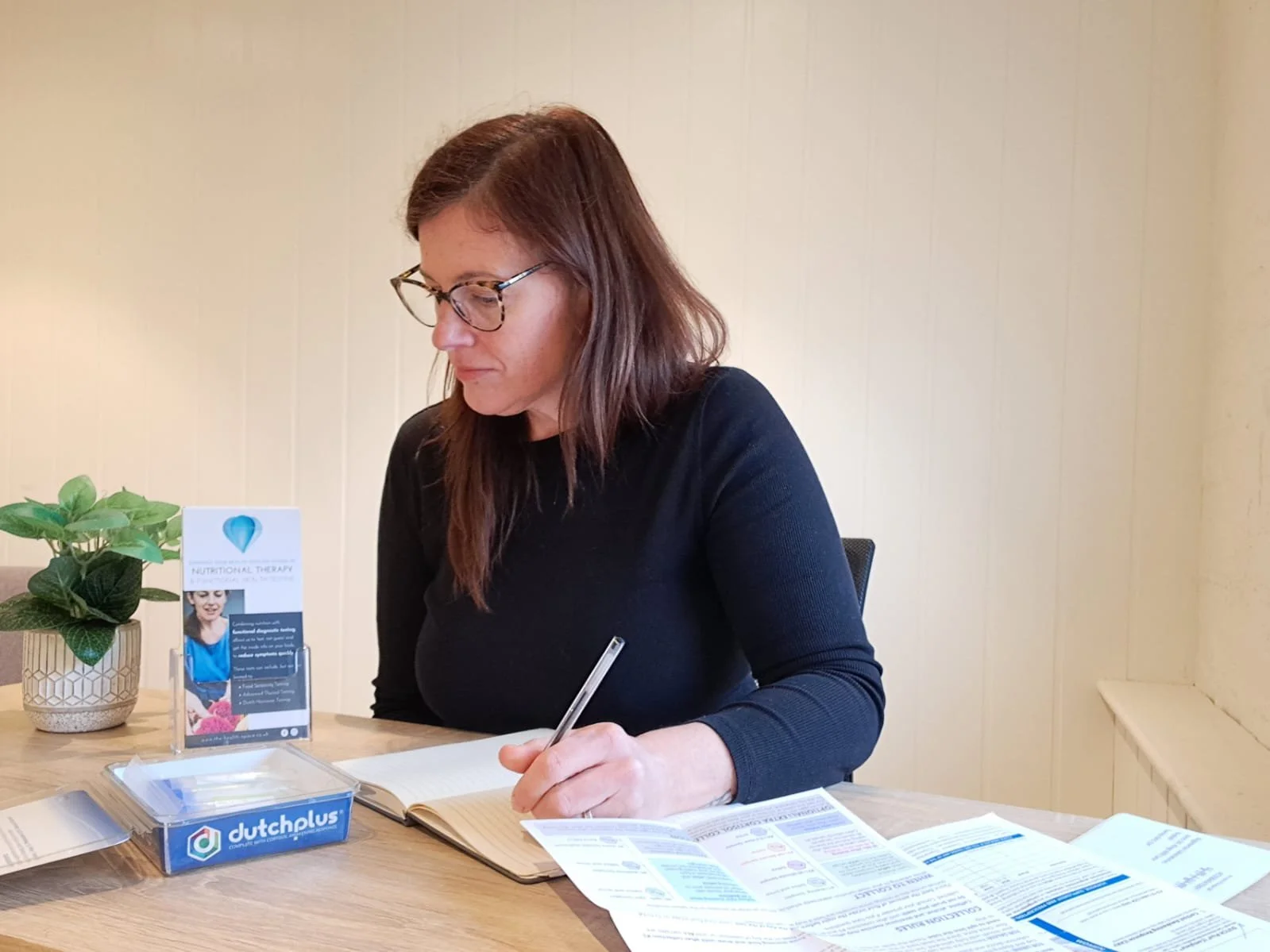 Woman with brown hair and glasses sitting at table, writing in notebook, with pamphlets and plant nearby.