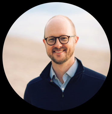 A man with glasses, a beard, and short hair smiling outdoors, wearing a navy blue jacket over a light blue shirt.