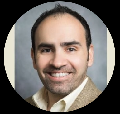 Headshot of a smiling man with dark hair, wearing a light-colored shirt and beige blazer, against a gray background.