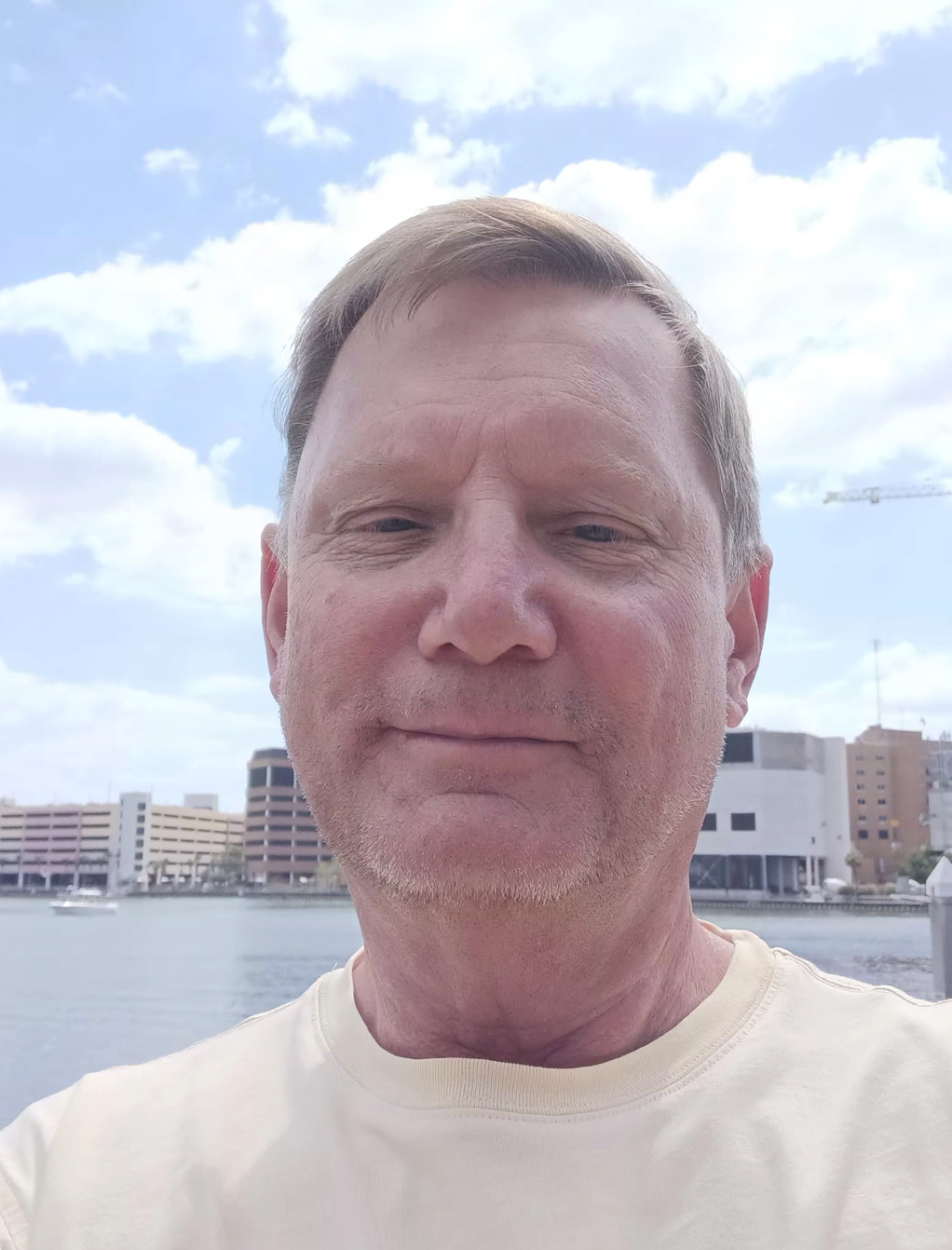 A man taking a selfie outdoors near water with buildings and a cloudy sky in the background.