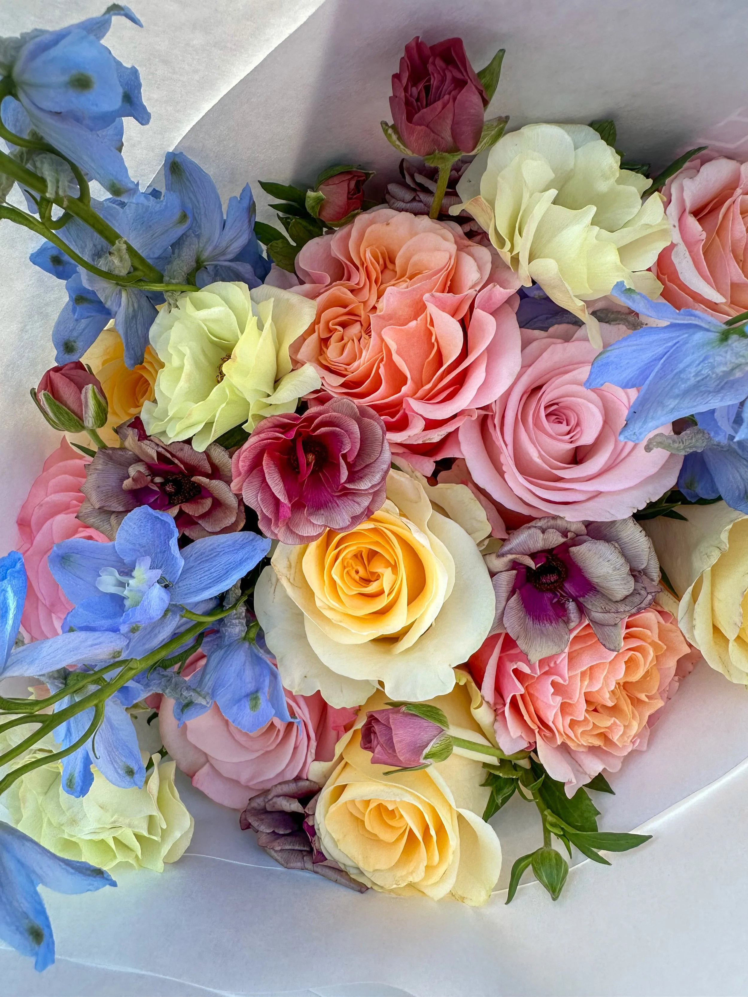 A bouquet of mixed pastel-colored flowers including roses, delphiniums, and ranunculus, wrapped in white paper.