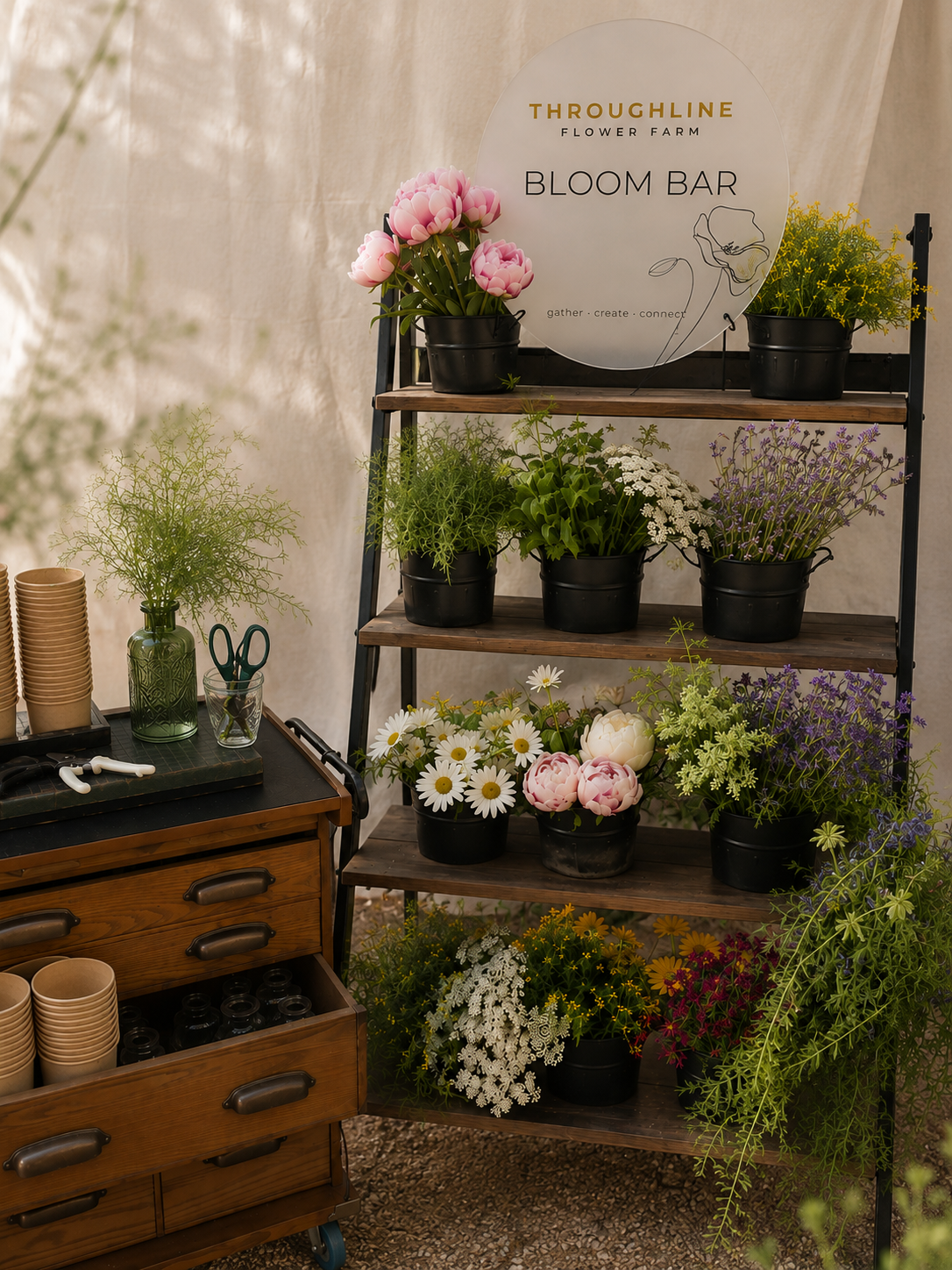 complete Throughline bloom bar outdoors — tiered wooden shelving, black buckets, frosted round sign, vintage drawer cart, umbrella