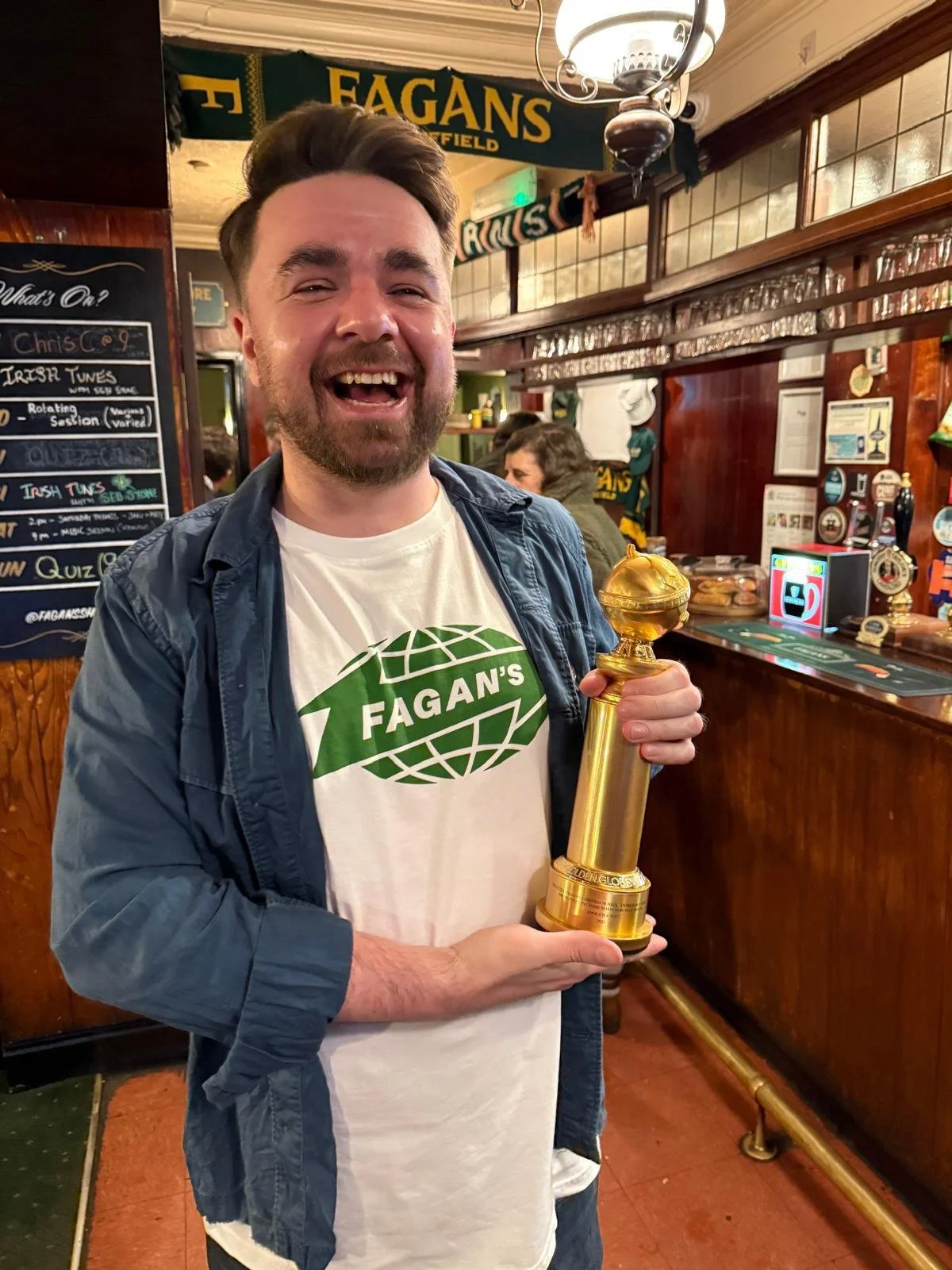 A man smiling and holding a gold trophy inside a pub named Fagan's. He is wearing a white T-shirt with a green logo and a blue jacket. The pub has wooden decor and a chalkboard menu behind him.