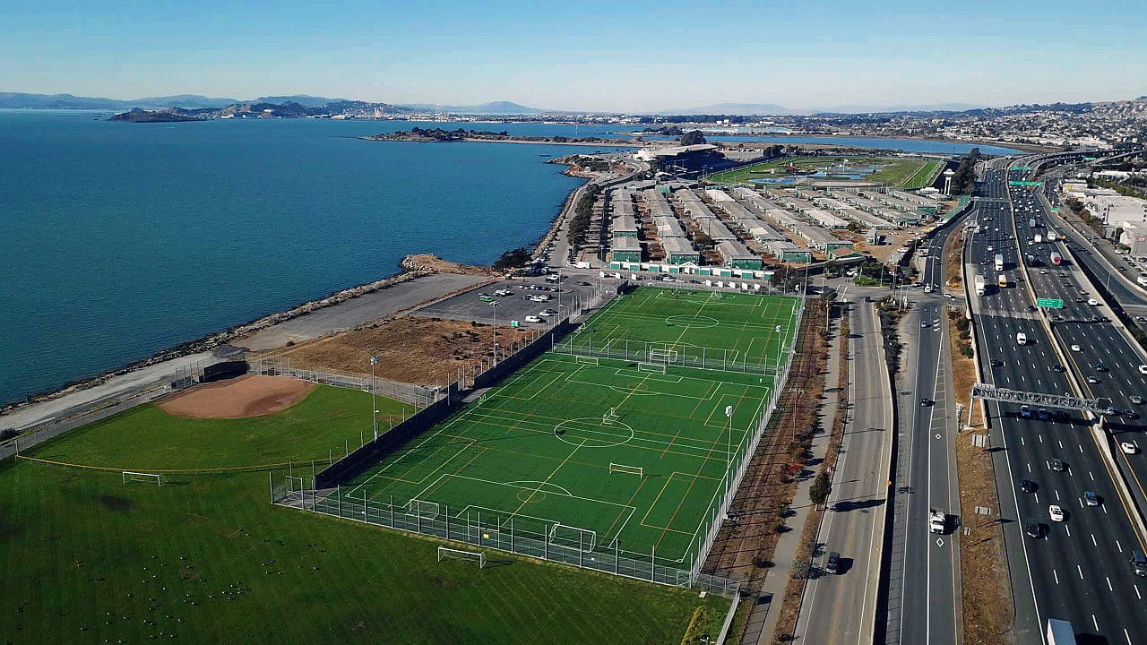 Aerial view of Gilman Fields at Tom Bates Regional Sports Complex in Berkeley, California, showing multiple turf soccer fields along the San Francisco Bay.