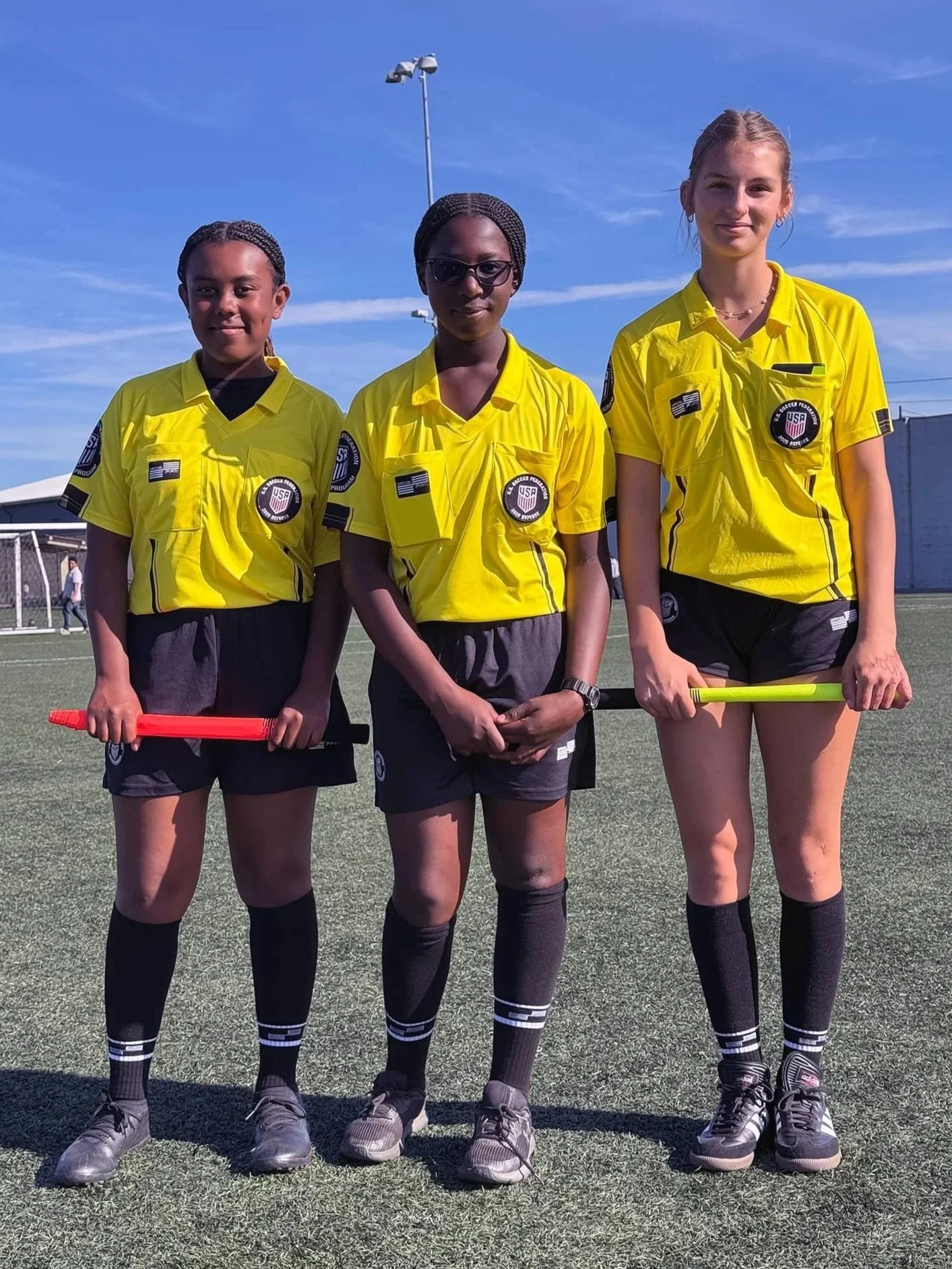 Female youth soccer referees pose on the field wearing official referee uniforms at an Eastshore Alliance FC match, highlighting referee education and leadership opportunities.
