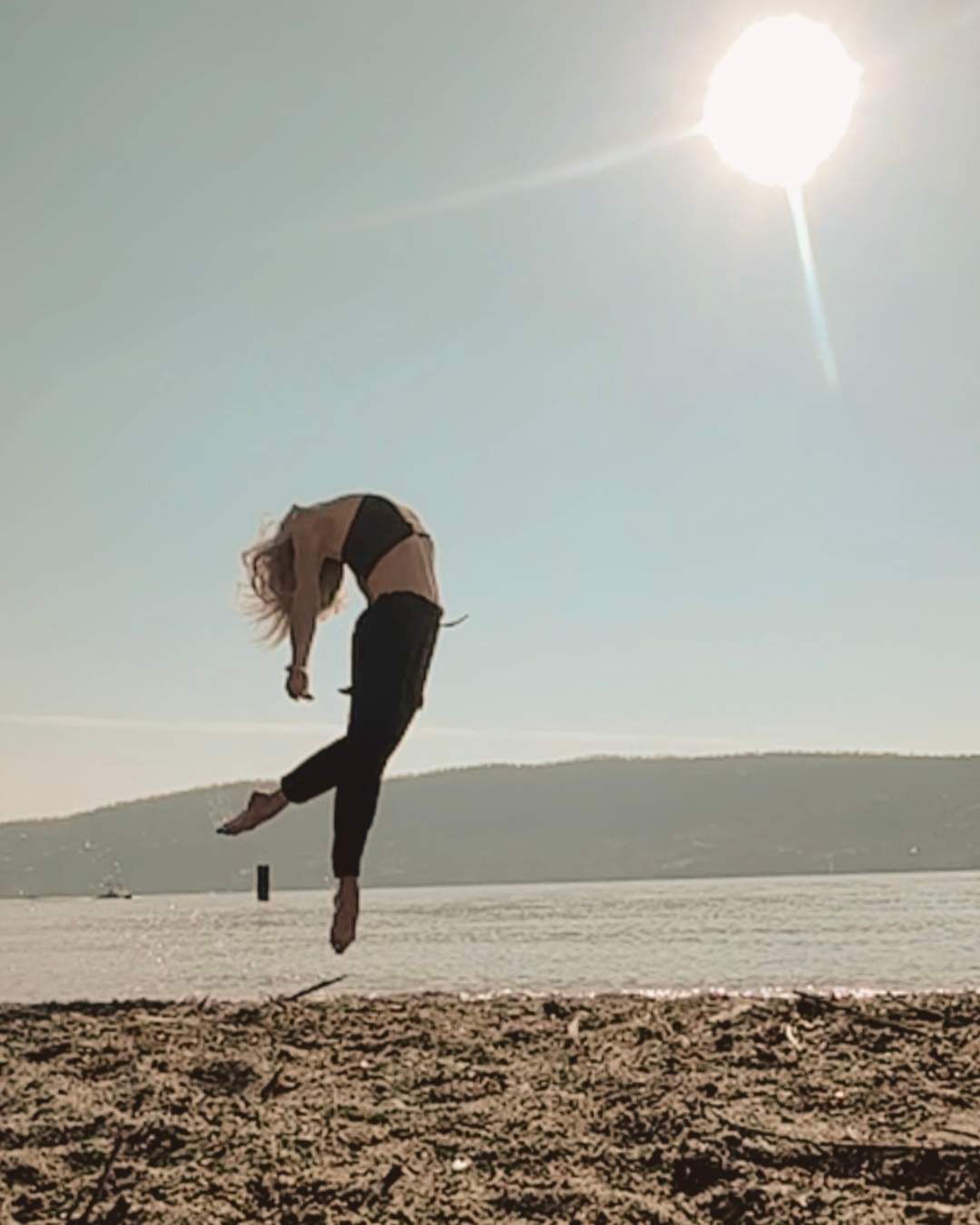 Person performing a dance jump on a beach with the sun shining brightly in the sky.