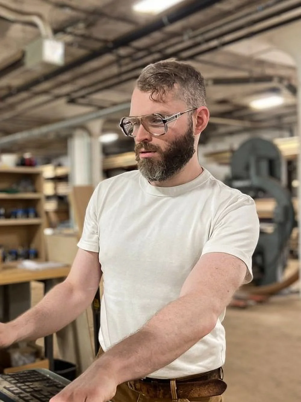 A person with glasses and a beard working in a woodworking shop, surrounded by wood and woodworking tools.