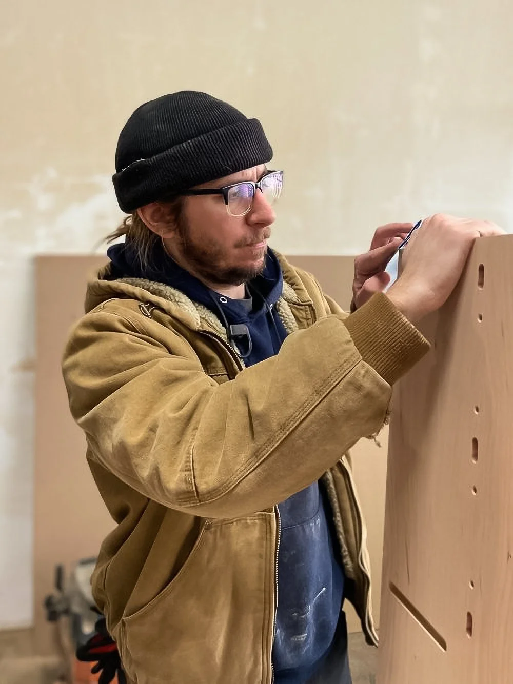 Man wearing glasses, a black beanie, a tan jacket, with a hoodie underneath, standing and marking a piece of light-colored wood with a pencil.