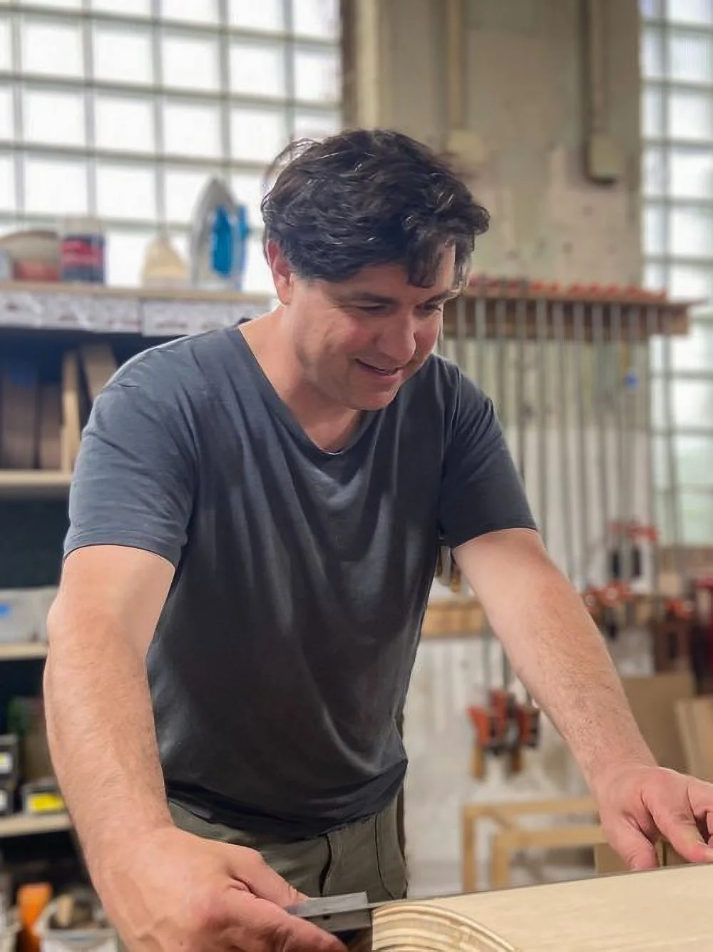A man is working on a woodworking project in a workshop, smiling while holding a tool and standing over a piece of wood.