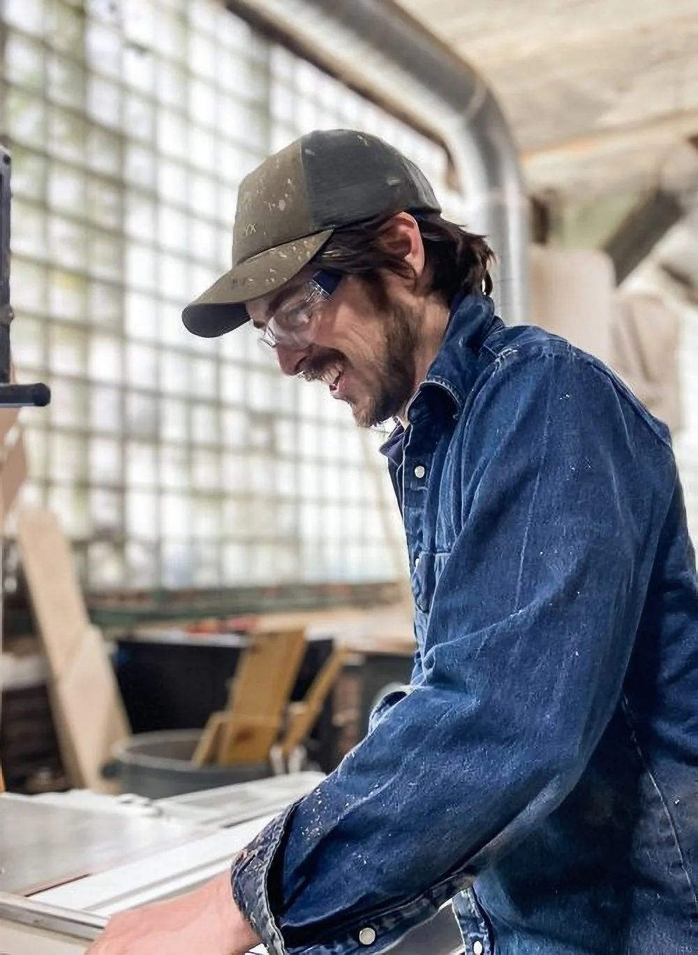 A man wearing a green cap, safety glasses, and a blue denim jacket, smiling while working with wood in a woodworking shop.