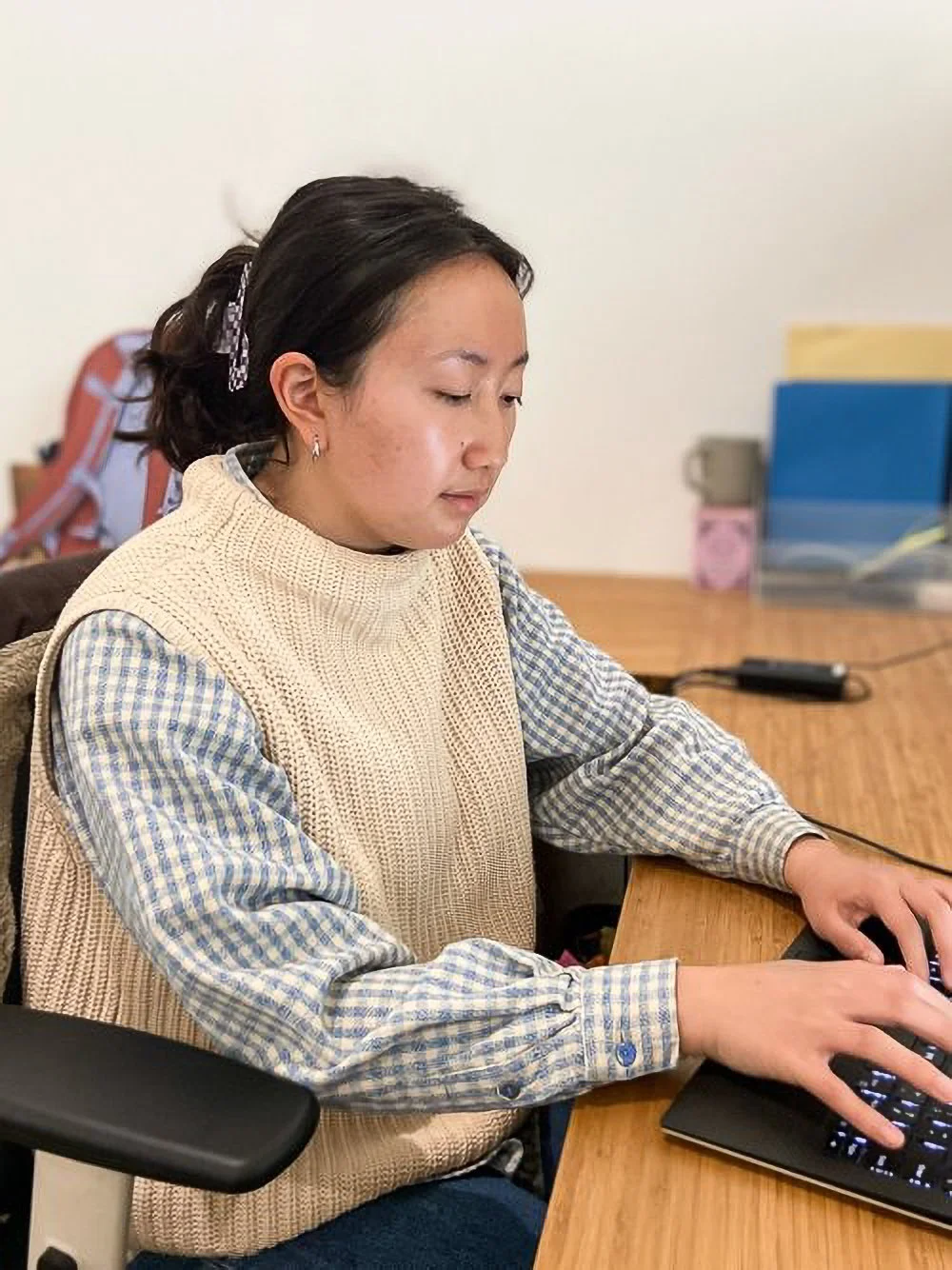 A woman with dark hair, tied back with a checkered hair clip, wearing a beige knitted vest over a blue and white checkered shirt, working on a computer at a wooden desk.