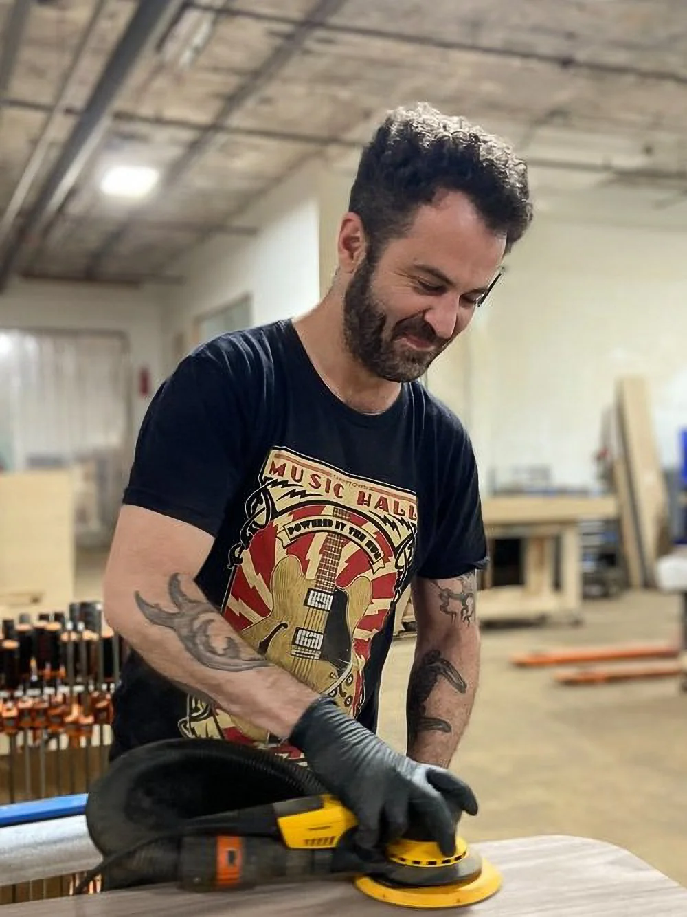 A man with tattoos on his arms using a handheld power sander on a piece of wood in a workshop.