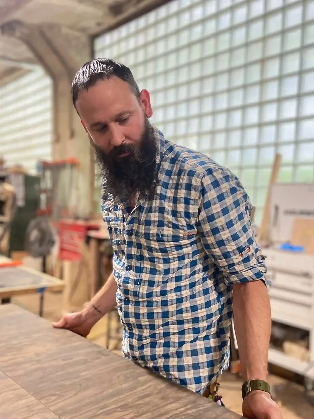 A man with a beard wearing a blue and white checkered shirt, leaning over a wooden work surface in a workshop or woodworking space.