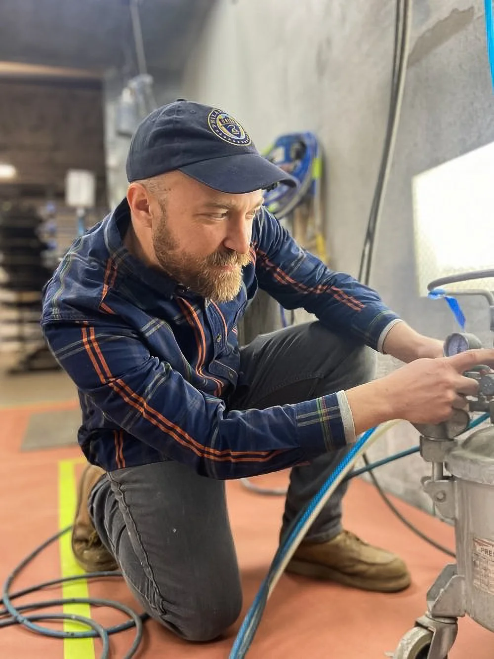 A man with a beard wearing a navy baseball cap and plaid shirt, kneeling on one knee, working with tools on a machine or equipment in an industrial or workshop setting.