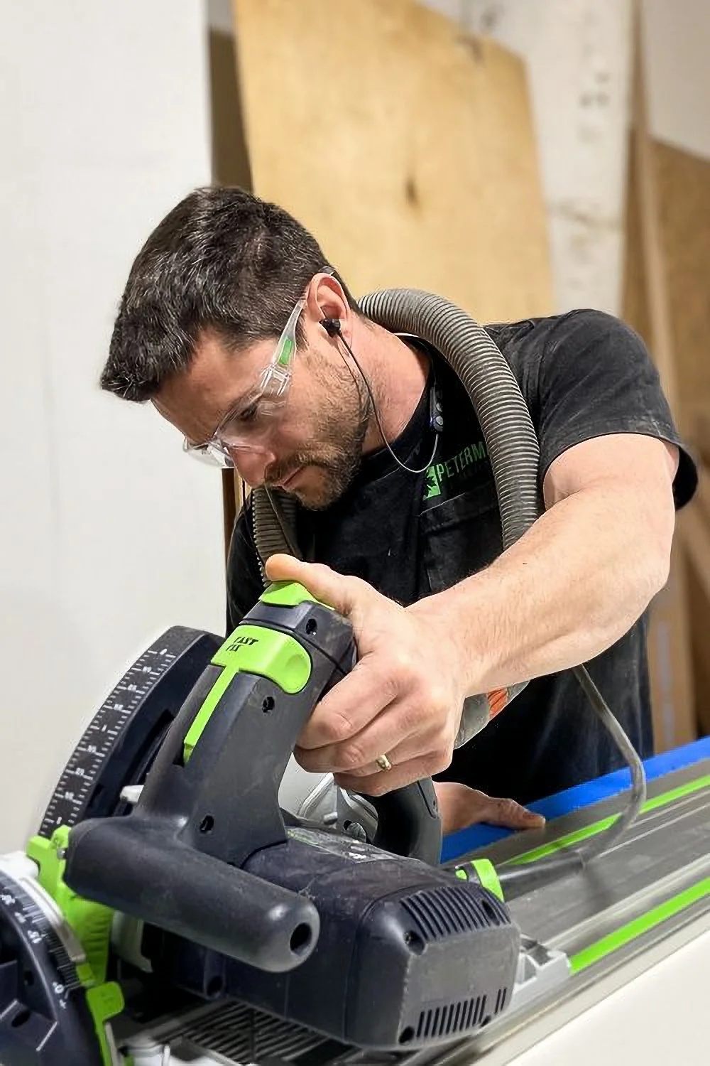 A man wearing safety glasses and ear protection is using a green and black power tool, possibly a saw, on a workbench in a workshop.