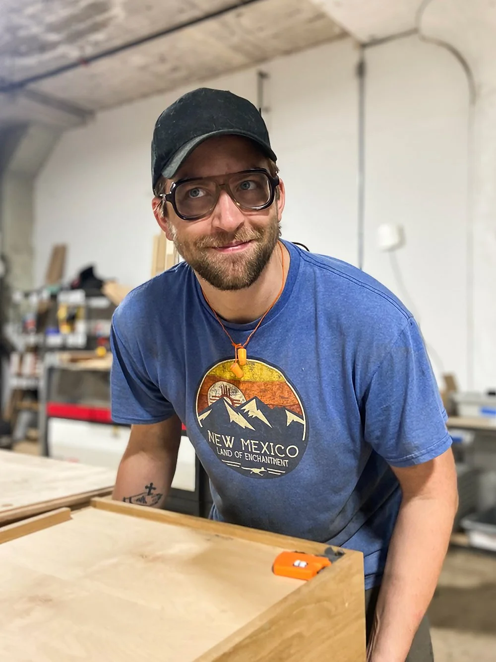 A man with a beard, glasses, and a black cap working in a woodworking shop, leaning over a wooden table with a tape measure on it, wearing a blue t-shirt that says 'New Mexico Land of Enchantment'.