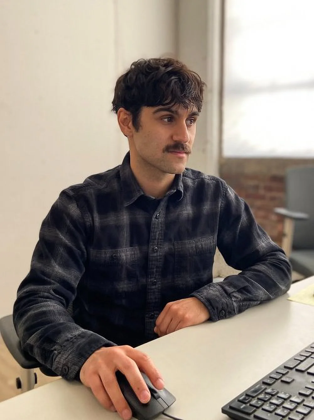 A young man with dark, curly hair and a mustache, wearing a black plaid shirt, sitting at a desk with a computer mouse in his right hand and a keyboard in front of him, in a room with a brick wall and a window with blinds in the background.